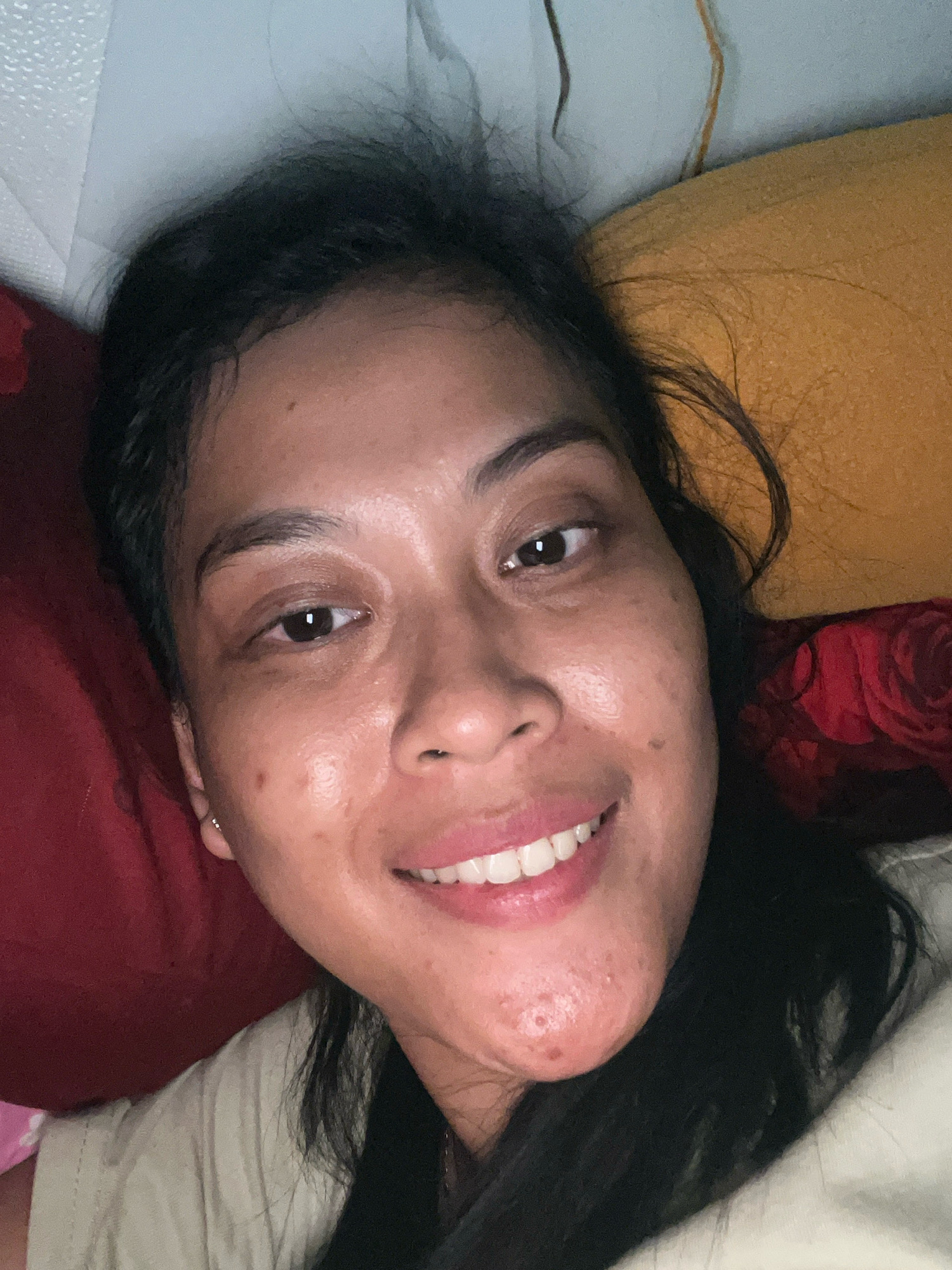 A close-up, overhead shot shows a woman lying down, her head resting on a pillow or cushion, with her face turned towards the camera. She has dark hair that is slightly messy and falls around her face. Her eyes are open and looking at the camera. Her mouth is open in a smile, revealing white teeth. Her skin appears to have some texture and blemishes, particularly on her chin and cheeks. She is wearing a light-colored shirt. Behind her, pillows or cushions in red and yellow are visible. The background is out of focus, but suggests an indoor setting. The image is grainy and appears to be taken in low light conditions. The woman is likely in her home in Kecamatan Pati, Indonesia, based on the provided context.