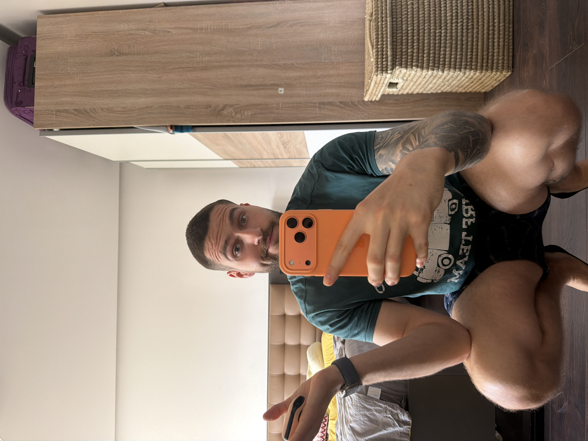 The image is a selfie taken in a room with a wardrobe, a wicker basket, and a bed with a headboard. The person taking the selfie is holding an orange phone and wearing a dark green t-shirt, dark shorts, and a watch on his left wrist. He has tattoos on his left arm and leg. The background includes a white wall, a wardrobe with light brown wooden doors, a wicker basket on top of the wardrobe, a bed with a light brown headboard, and gray sheets. The room appears to be a bedroom. No location data is provided.
