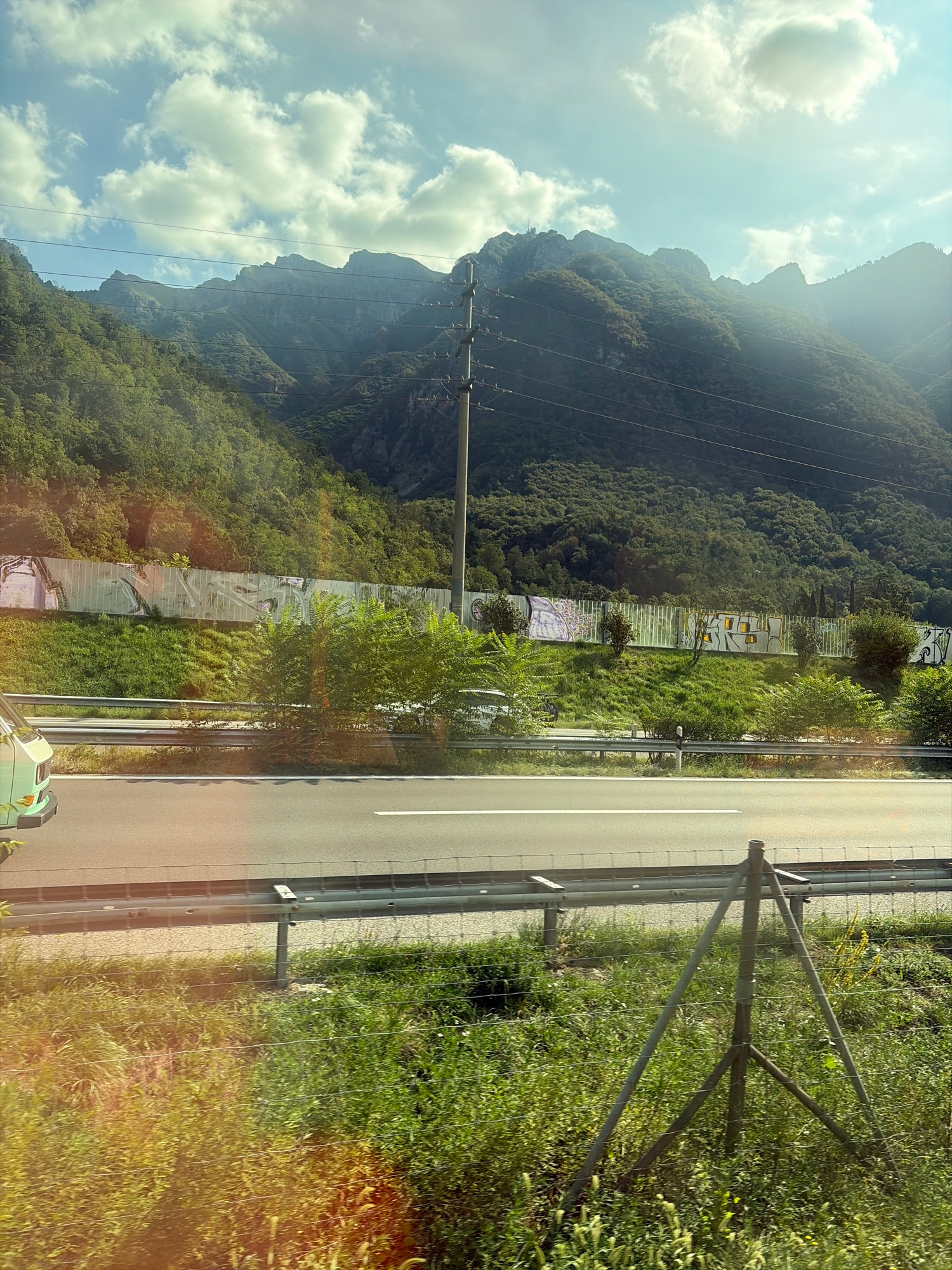 The image shows a view from a moving vehicle on a highway in Val Mara, Switzerland. In the foreground, a green van is partially visible on the left side of the road. The highway stretches across the mid-ground, with a metal guardrail separating it from a grassy embankment. This embankment is topped with a metal fence adorned with graffiti, and further beyond, a dense, green forest covers a steep mountainside. The upper portion of the image is dominated by a bright, blue sky filled with fluffy white clouds, and in the distance, the rugged peaks of the mountains are silhouetted against the sky. A utility pole with electrical wires is positioned on the right side of the frame, running parallel to the road. The overall scene depicts a typical roadside view of the mountainous landscape of Val Mara.
