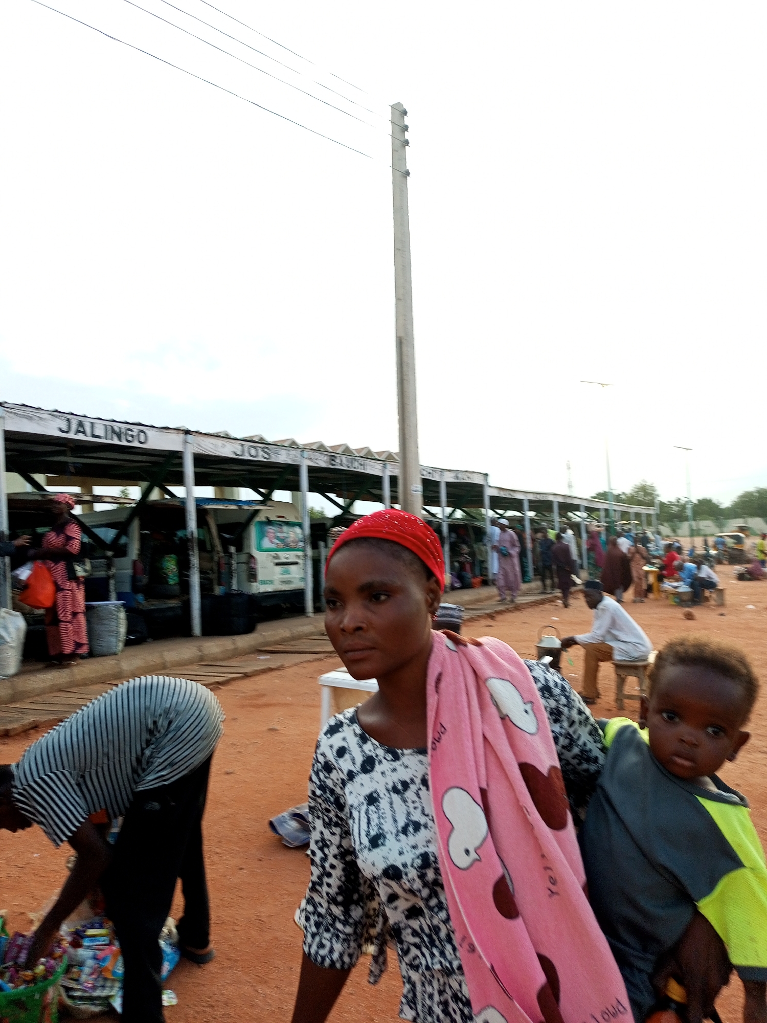 The image depicts a bustling outdoor marketplace in Gombe, Nigeria. In the foreground, a woman wearing a red headscarf and a black and white patterned dress is prominently featured, holding a pink blanket with white heart and bird patterns. To her right, a young child with dark skin is being held by someone, looking towards the camera. To the left, a person wearing a striped shirt and black pants is bent over a basket filled with various items. In the background, a covered market structure with signage indicating "JALINGO" and "JOS" is visible, with several vehicles and a crowd of people milling about. The ground is covered in reddish-brown dirt, and overhead, power lines crisscross against a bright, overcast sky. The overall scene suggests a lively commercial hub where people are engaged in shopping, travel, and daily activities.