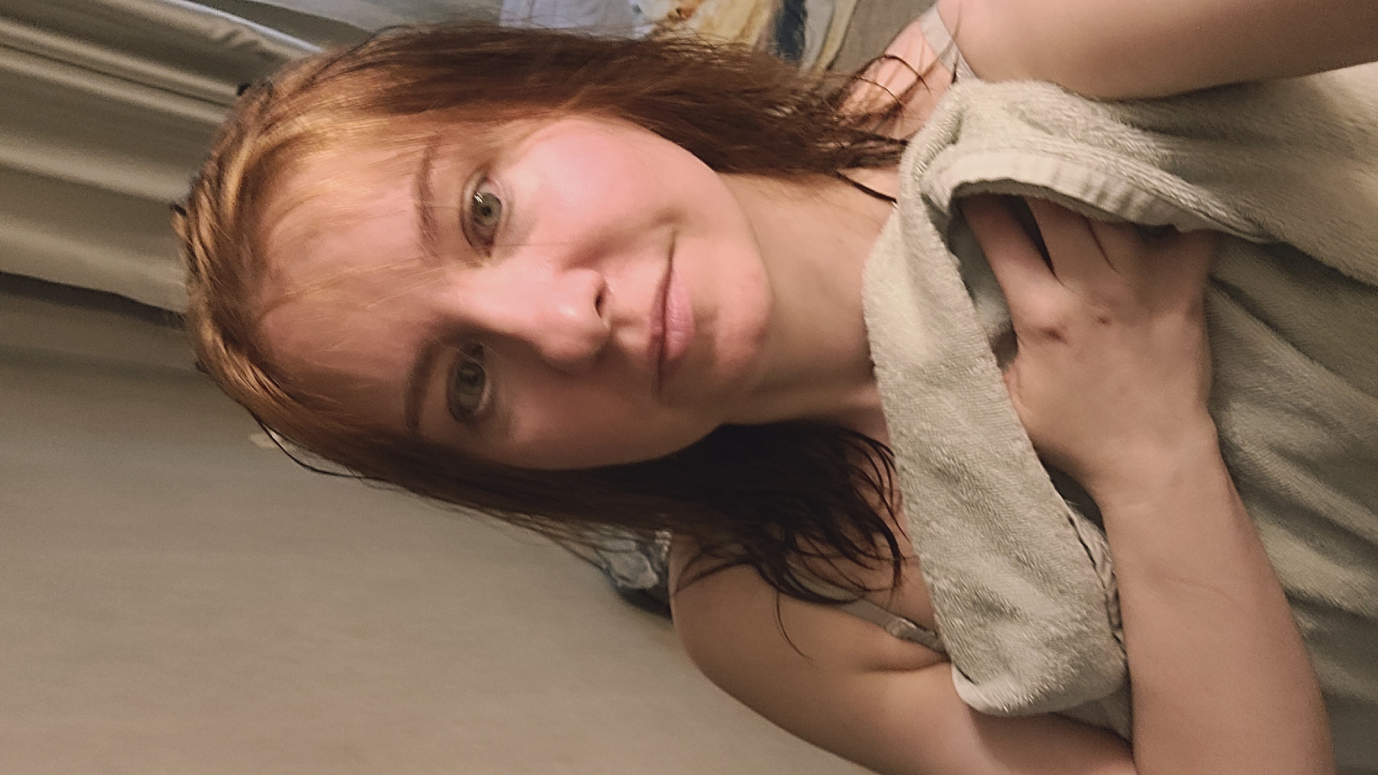 A woman with auburn hair is wrapped in a light green towel, looking at the camera. Her hair is wet and styled in loose waves, with some strands falling across her forehead. She is wearing a bra that is partially visible beneath the towel. The background appears to be a domestic setting, with a plain wall and curtains visible. The lighting is soft and ambient, suggesting an indoor environment. The woman's expression is neutral, and her eyes are looking directly at the viewer. Her right hand is tucked into the towel, with her fingers slightly exposed. The overall impression is a casual, intimate portrait.