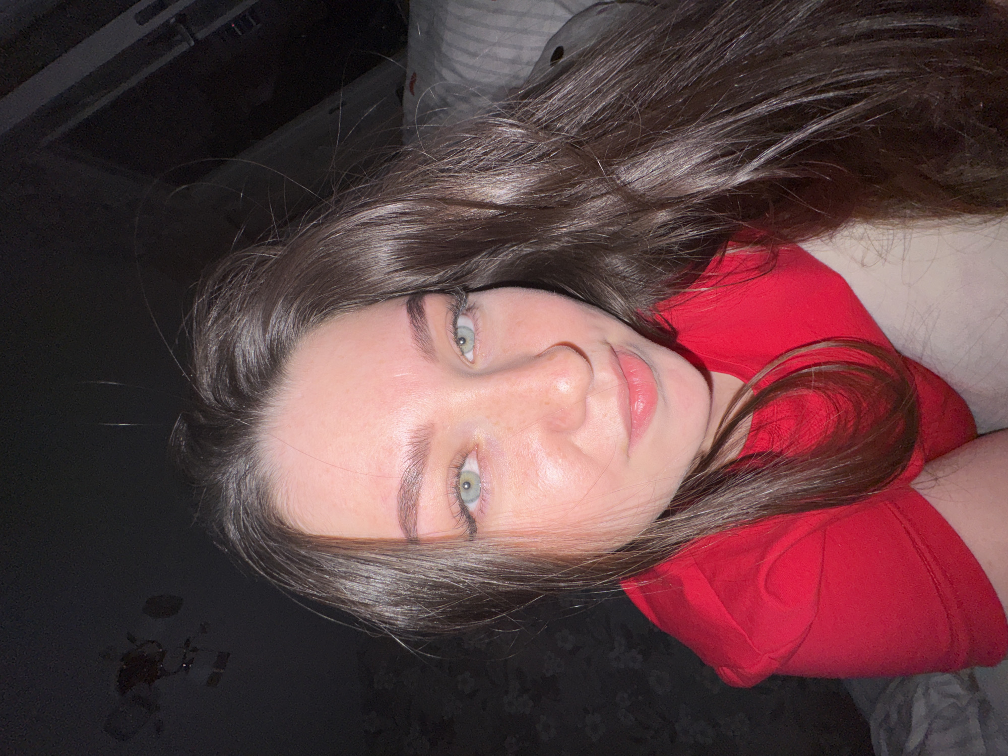 A medium close-up, portrait-style photograph shows a young woman lying down and looking directly at the viewer. She has long, wavy dark brown hair, which is spread out around her head and across her shoulders. Her eyes are a striking light green or hazel, and her fair skin shows subtle freckles on her nose and cheeks. Her lips are slightly parted, showing a hint of a light pink lip product. She is wearing a bright red top or t-shirt. The woman's expression is calm and neutral, with a gentle gaze.
The perspective is from above, suggesting she might be lying on her back, possibly on a bed or soft surface, parts of which are visible as blurred, light-colored fabric (potentially a pillow or duvet) in the upper right background. The background is predominantly dark, indicating low or dim artificial lighting, possibly suggesting an indoor setting like a bedroom during the evening or night. There are no specific objects or features in the background to provide distinct location cues. No visible text is present in the image. The provided context states the photograph was taken in Perm, Russia.