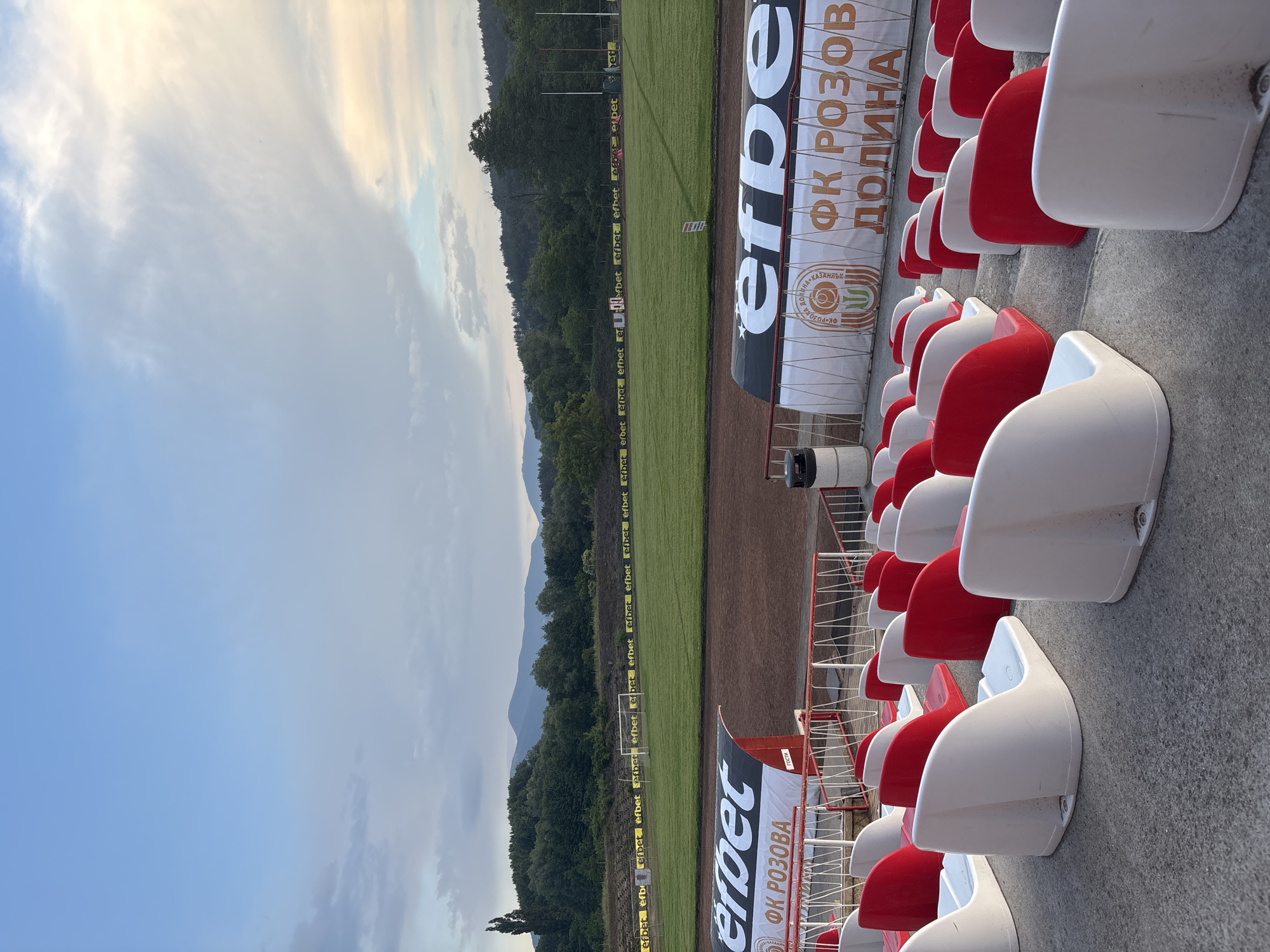 This image depicts a football stadium, likely in Kazanlak, Bulgaria, as indicated by visible text. The scene is captured during what appears to be late afternoon or early evening, characterized by soft, diffused sunlight and a sky with scattered white and grey clouds, blending into a light blue. No people are visible, suggesting the stadium is empty, perhaps before or after a game or training session.
The foreground features rows of tiered stadium seating, with individual plastic seats alternating in red and white. These seats are arranged on concrete steps, providing a perspective looking towards the pitch. In the midground, a neatly maintained green grass football pitch is visible, bordered by a reddish-brown track. Around the perimeter of the pitch, numerous advertising hoardings prominently display the brand "efbet" multiple times. A white goalpost can be seen on the far side of the pitch.
Beyond the pitch, a dense line of green trees forms a natural barrier, with distant mountains rising softly against the horizon, partially obscured by the atmospheric haze. Banners attached to the stadium railings near the seating areas feature Cyrillic text. A large banner clearly reads "ФК РОЗОВА ДОЛИНА" (FK Rozova Dolina), identifying the local football club. Other text includes "КАЗАНЛЪК" (Kazanlak), confirming the location context, and "гости" (guests) on a smaller sign. The overall impression is one of a peaceful, well-maintained sports facility awaiting or having just completed activity.