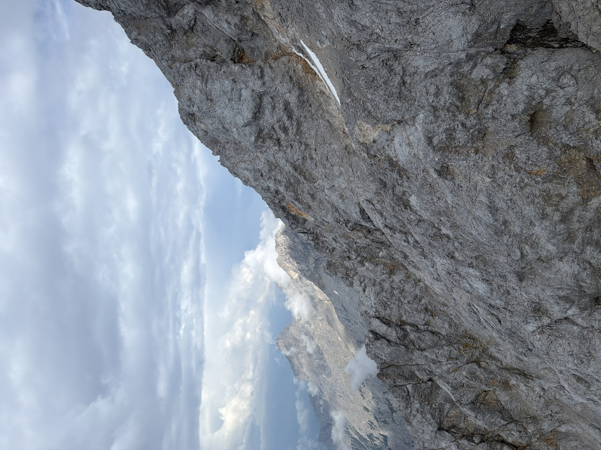 This is a landscape photograph taken from a high vantage point, looking up at a sheer, gray rock face. The rock is rough and textured, with visible cracks and variations in color, suggesting a natural geological formation. A thin strip of white, likely snow or ice, is visible on the rock face.
Below and to the left of the rock face, the sky is filled with dramatic, puffy clouds. Gaps in the clouds reveal glimpses of distant, rugged mountain peaks, some of which are also shrouded in mist or low-lying clouds. The overall impression is one of a dramatic, mountainous environment.
The lighting suggests it is daytime, possibly late morning or early afternoon, given the brightness of the sky and the way light plays on the rock. The weather appears to be variable, with both clear sky and cloud cover, and the presence of snow/ice indicates a high altitude or a cool climate.
The image conveys a sense of awe and perhaps a touch of the formidable power of nature. The perspective is from someone looking up at the immense scale of the rock and the vastness of the sky, suggesting a hiker, climber, or someone observing the grandeur of the Alps. The location context of Grainau, Germany, places this scene in the Bavarian Alps. There is no visible text in the image.