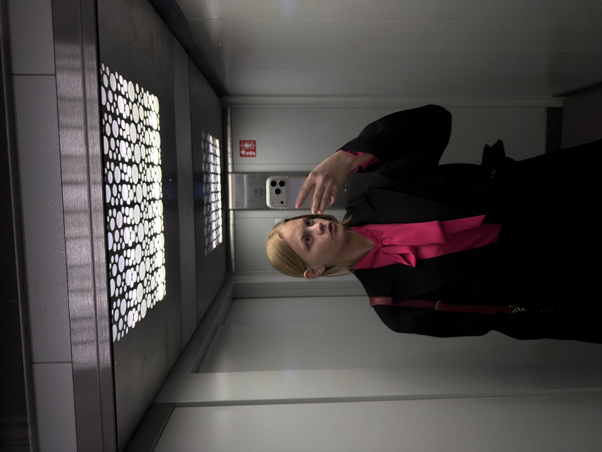 A woman is taking a selfie inside an elevator. The elevator's doors have a decorative pattern of circular cutouts. The woman is wearing a black coat over a pink top. A red purse strap is visible over her shoulder. The elevator interior is a neutral gray. A sign with a restroom symbol is visible on the back wall of the elevator. It's not possible to confirm that the image was taken in Izhevsk, Russia.