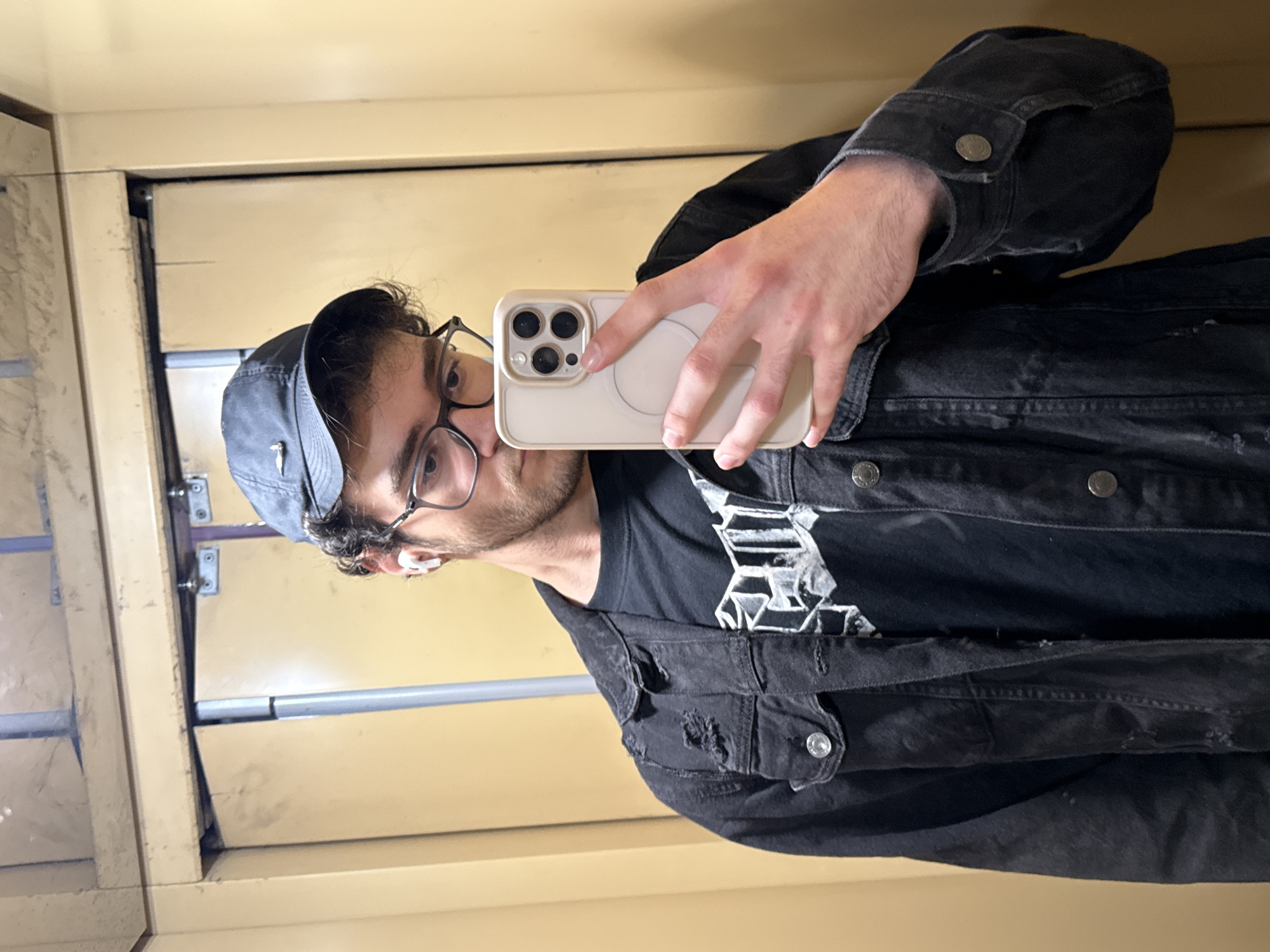 The image captures a selfie of a man inside what appears to be an old elevator or a utilitarian room. He is seen from the chest up, looking directly at his reflection while holding a light-colored smartphone to take the picture. The man wears a dark baseball cap, clear-framed glasses, a black t-shirt with a white, abstract graphic design, and a distressed black denim jacket. White wireless earbuds are visible in his ears. The smartphone, held in a light-colored case, clearly shows three large camera lenses on its back.
The background features pale yellow-beige walls that are visibly worn and scratched, indicating an aged interior. The walls are constructed from panels with distinct vertical seams. A horizontal metal bar or rail runs across the background, and various metallic fixtures, possibly hinges or bolts, are discernible, reinforcing the impression of an enclosed, functional space like an old elevator car. A faint, dark logo, resembling the Nike swoosh, is visible on his cap. The graphic on his t-shirt consists of angular, geometric shapes in white. No other specific text is clearly decipherable. The lighting suggests an indoor setting, typical for an enclosed space.