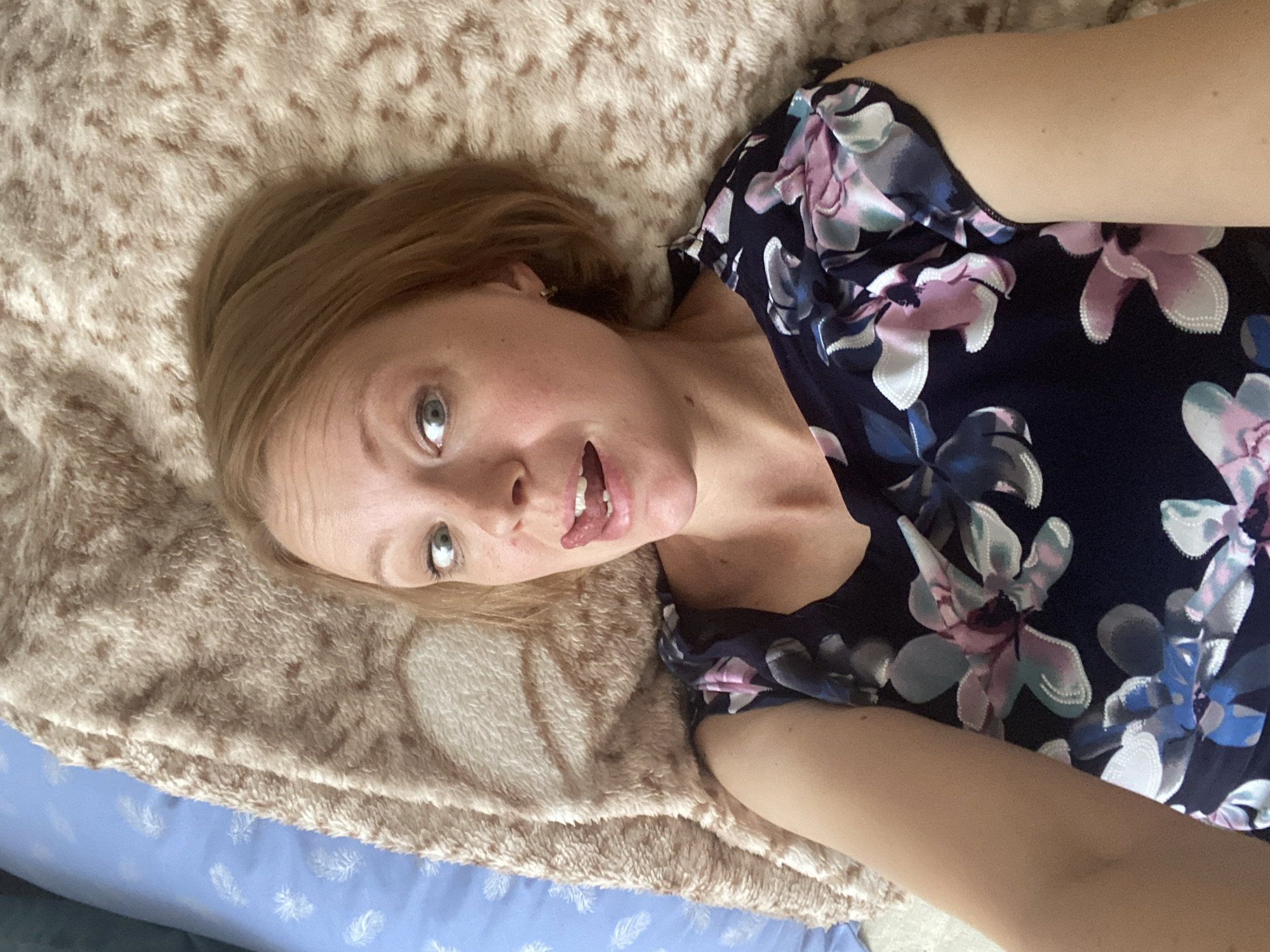 A woman with blonde hair and light blue eyes is lying on her back on a brown, fuzzy blanket. She is wearing a dark blue dress with a floral pattern of pink, white, and blue flowers. Her mouth is open and her tongue is sticking out. She is looking directly at the camera. The background is a blue pillow with a white feather pattern. The image was taken in Maykop, Russia.