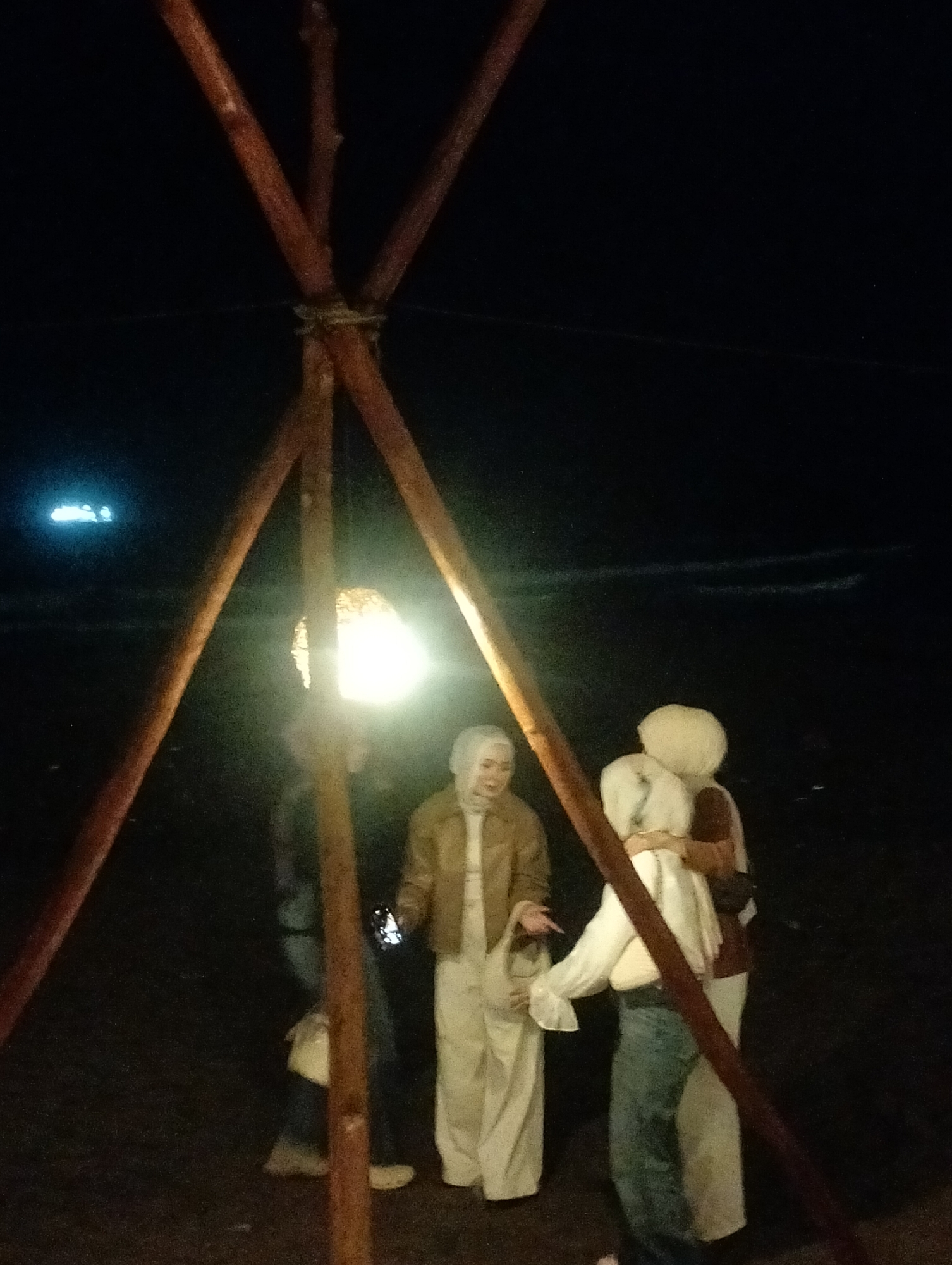The image captures a nighttime scene in what appears to be an outdoor setting in Keudekruenggeukueh, Indonesia. A group of four individuals, predominantly women, are gathered around a rustic wooden tripod structure from which a bright light source hangs, illuminating the immediate area.
The subjects include three women clearly visible in the foreground. One woman, wearing a light brown jacket and white attire including a hijab, stands near the center, looking towards the right and smiling. Another woman to her right, also in a hijab and light-colored clothing, holds what appears to be a small child or object wrapped in white fabric, embracing it. A third person, partially obscured behind the light and wearing jeans and a darker top, is visible on the far left. The fourth person, positioned behind the tripod and to the right, is mostly obscured but appears to be wearing a hijab and light clothing, possibly looking at a phone held in their hand, which is faintly lit.
The activity taking place seems to be a casual social gathering or an outdoor evening event. The people are standing close together, interacting, and one person is looking at a phone, suggesting a relaxed atmosphere.
Notable details include the time of day, which is clearly night, with the only significant illumination coming from the lamp suspended from the wooden tripod. The background is almost entirely dark, with distant, faint blue lights visible on the far left horizon, possibly indicating a coastline or distant structures. The wooden tripod itself is made of rough-hewn branches tied together at the top, suggesting a makeshift or natural design. The visible faces appear relaxed, with at least one person smiling, conveying a sense of ease among the group. There is no visible text in the image.