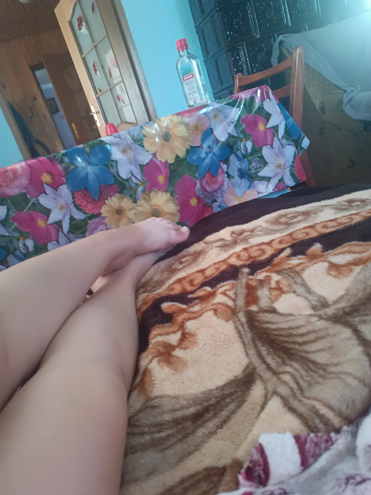 The image shows a pair of legs and feet resting on a blanket with a brown and tan patterned design. The legs are bare and appear to be light-skinned. The blanket is folded and draped over what seems to be a couch or bed. In the background, a table is visible, covered with a colorful tablecloth featuring a floral pattern. A bottle of liquid with a red cap is placed on the table. Further in the background, a wooden chair, a wooden door, and a dark-colored tiled stove are discernible. The overall lighting suggests an indoor setting, possibly a room in a house in Sokoliv, Ukraine.