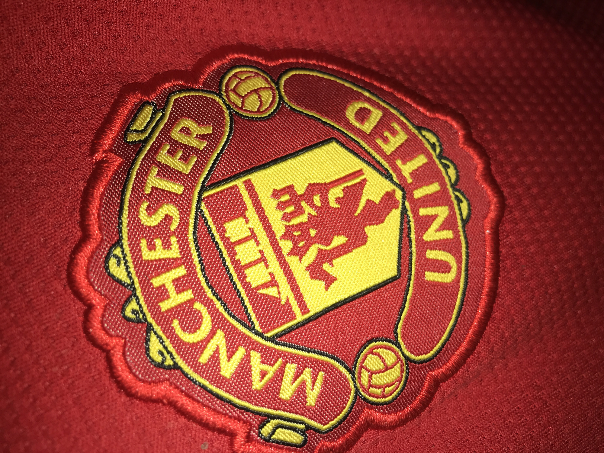 A close-up shot features an embroidered Manchester United football club badge prominently displayed on a vibrant red fabric, likely a sports jersey or similar apparel. The badge is intricately detailed, primarily in red and yellow, and stands out with its raised, textured embroidery.
The central element of the badge is a yellow shield, emblazoned with the iconic red "red devil" figure. Above this, a stylized ship emblem is depicted on the top of the shield. Two classic yellow footballs are positioned on either side of the shield, near the top, adding to the sporting theme. The club's name, "MANCHESTER UNITED," is stitched in yellow capital letters with fine black outlines, curving around the lower perimeter of the badge. "MANCHESTER" is visible on the left arc, and "UNITED" on the right.
The entire emblem is framed by a distinct red and yellow stitched border that gives it a three-dimensional quality against the background. The red background fabric itself shows a subtle, slightly textured pattern, characteristic of modern sports fabrics.
The image is a still life of an object, with no specific activity or event taking place. There are no discernible cues for the time of day or weather. The detailed stitching highlights the craftsmanship of the badge. This item is presented within the location context of a New Stadium in Nigeria.