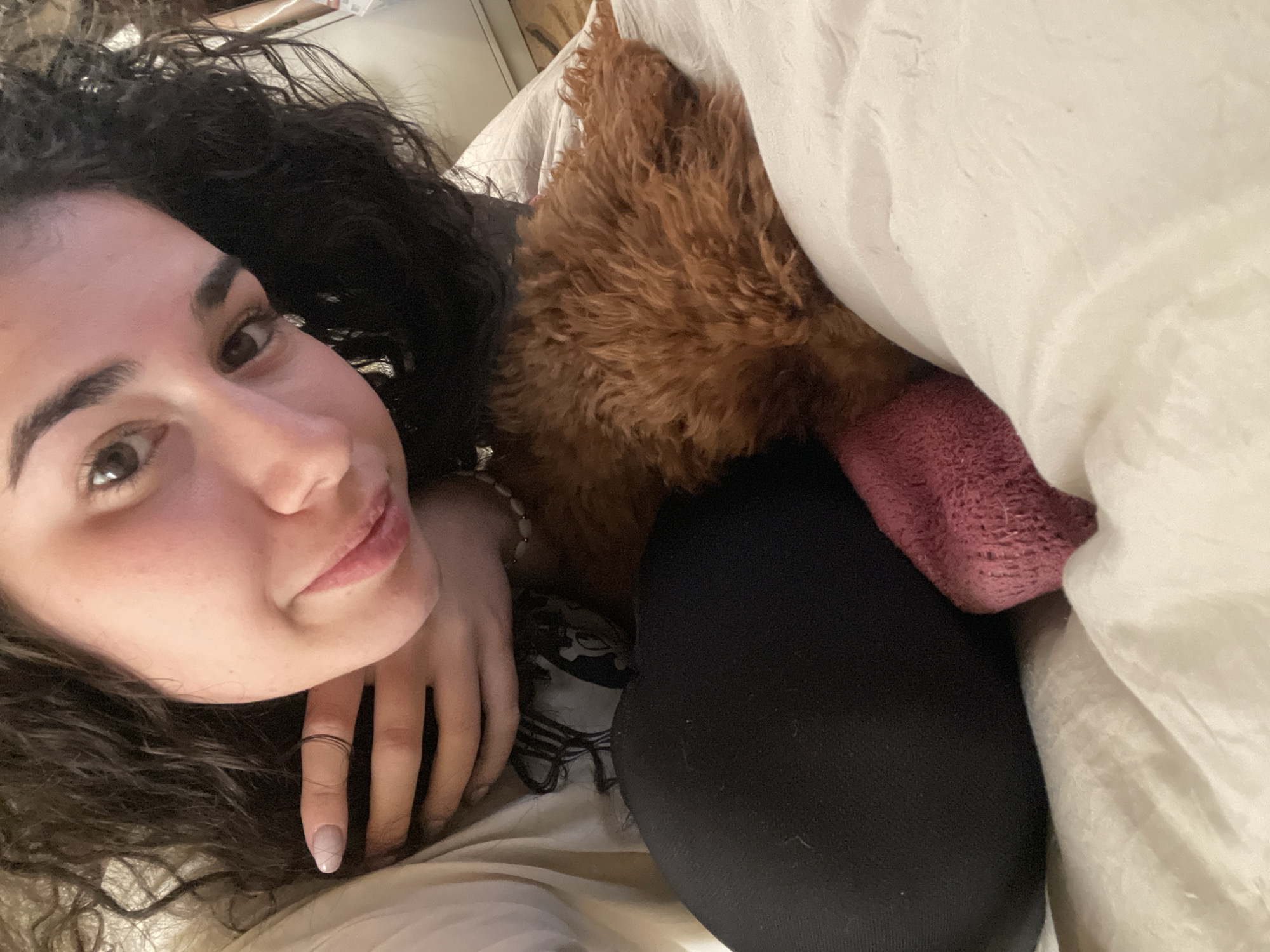 A woman with dark, curly hair is lying down, possibly in a bed. Her face is visible, and she has a ring on one of her fingers. A brown, furry stuffed animal with a pink end is next to her. The setting appears to be indoors on bedding. There is no visible evidence to place this photo in Sofia, Bulgaria.