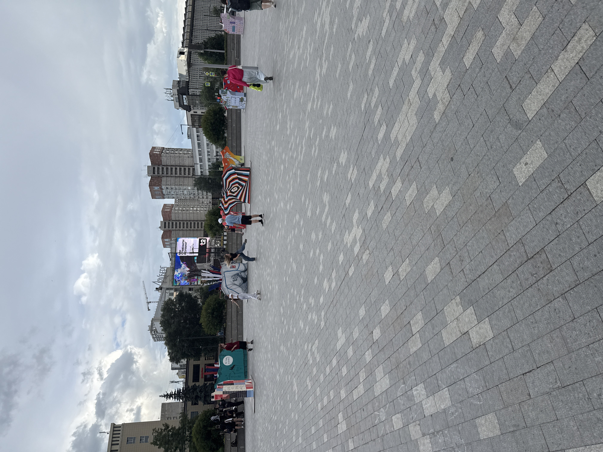 The image, taken in Perm, Russia, depicts a public square with a paved ground made of gray and light gray rectangular tiles. The scene is viewed from a high angle, with the ground filling the majority of the frame. In the background, a row of tall, modern buildings with many windows are visible, suggesting an urban environment.
Several people are scattered across the square. Some are standing, while others appear to be walking or engaged in activities. One person in a red jacket and light gray pants is holding a yellow bag. Another individual is wearing a white outfit and standing next to a large, colorful, patterned rug that is laid out on the ground. The rug features a circular design with concentric rings of various colors, including black, white, red, yellow, and blue. A few other people are seen in the distance.
In the background, a large electronic billboard displays bright imagery and text. A crane is also visible, indicating ongoing construction or development in the area. The sky is overcast with clouds, suggesting a cloudy day. The overall mood of the scene appears to be casual and public, with people enjoying the outdoor space. The presence of the billboard and modern buildings points to a contemporary urban setting.