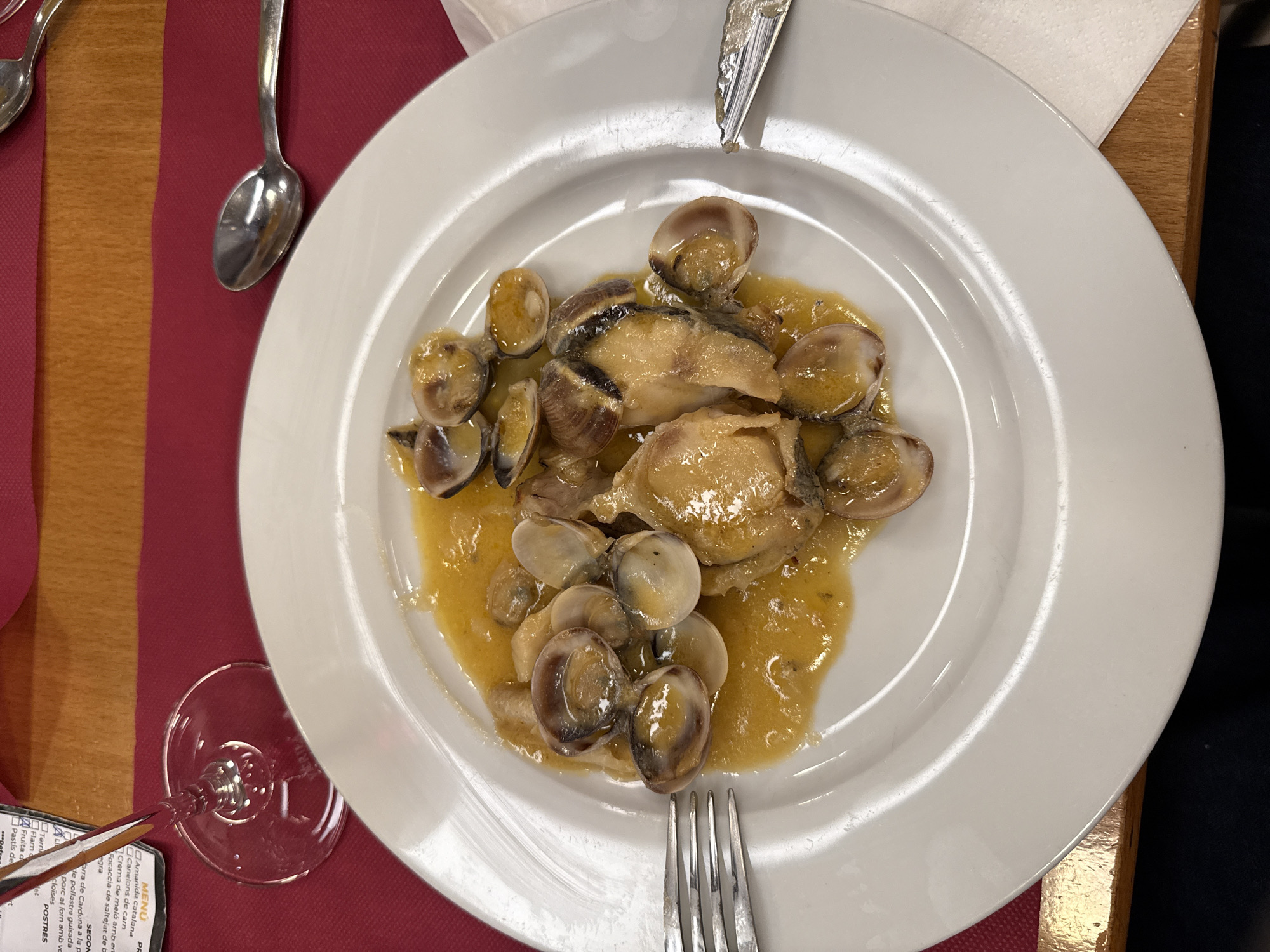 A top-down view shows a white plate filled with clams in a yellow sauce, interspersed with pieces of fish. The plate is placed on a red tablecloth with a wooden border. To the left, a wine glass is partially visible, and further to the left, a spoon rests on the tablecloth. A fork is positioned at the bottom right of the plate. In the background, a menu is visible with text in Catalan. The menu lists items such as "Amanida catalana," "Canelons de carn," and "cloïses." The presence of the menu suggests a dining setting, possibly a restaurant, in Cardona, Spain. The lighting indicates it's likely indoors, perhaps during the day or evening. The overall scene captures a close-up of a plated seafood dish with accompanying dining utensils and a menu.

--- OCR Start ---
AL
MENÚ
PR
Amanida catalana
Canelons de carn
Crema de meló amb en
Focaccia de saltejat de b
egra
SEGON
arra de Cardona a la p
de pollastre guisada
porc al forn amb ve
cloïses
Terri
Flam
Fruita d
Pastís de
POSTRES
--- OCR End ---