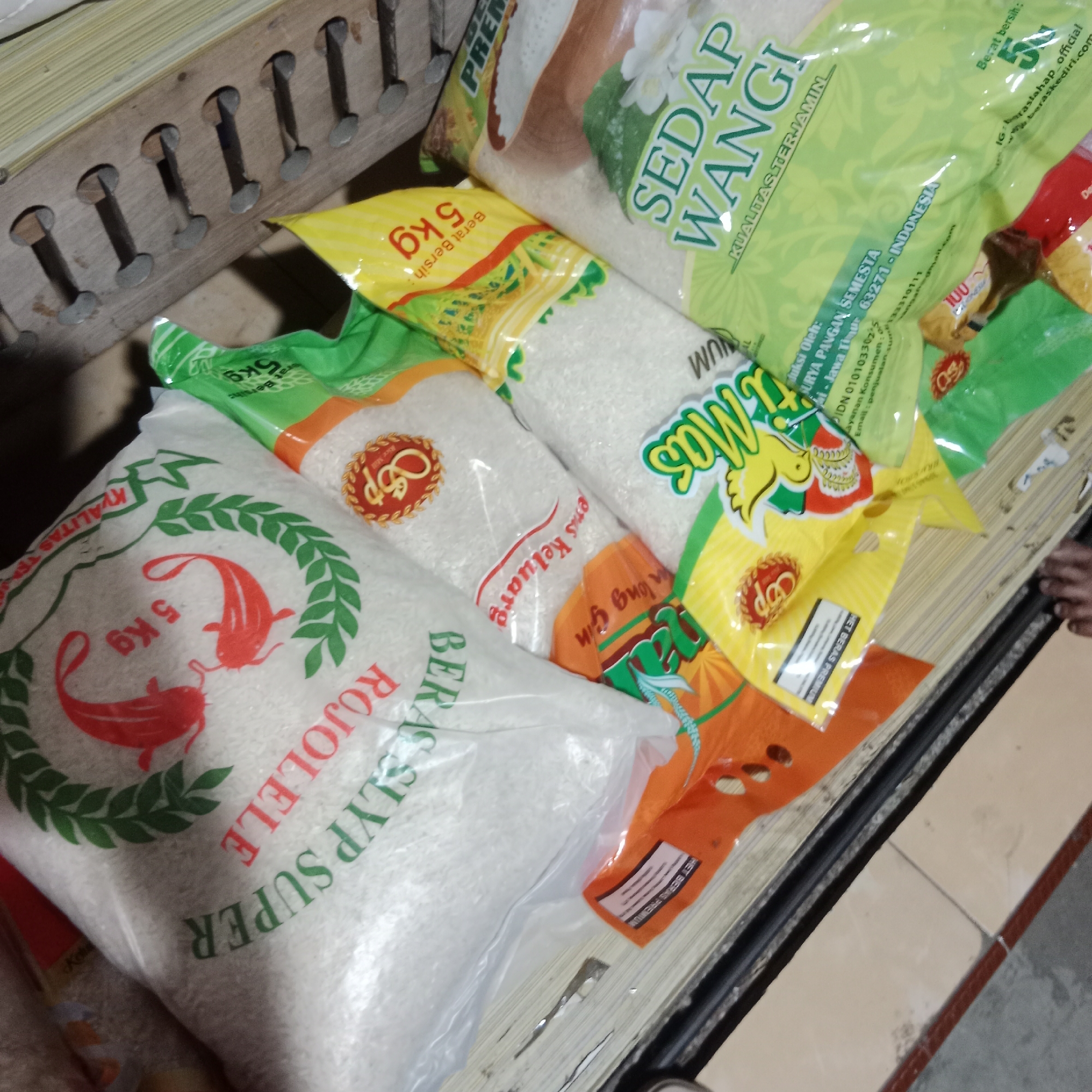 A pile of bagged rice is displayed on a wooden surface. The rice bags are of various brands and colors, including green, yellow, and white. Some bags have visible text in what appears to be Indonesian, such as "SEDAP WANGI" and "5kg". The rice bags are stacked and tilted, with some partially obscuring others. A wooden structure with metal fasteners is visible behind the rice bags. The lighting in the scene is dim, suggesting an indoor environment. The overall scene depicts a retail or storage area for rice products.