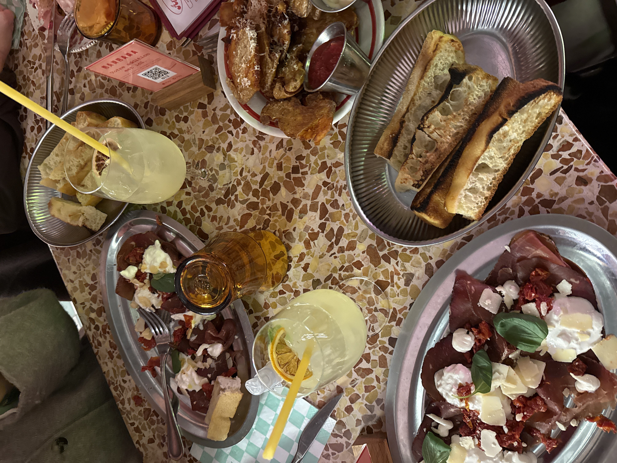 The image depicts a table laden with food and drinks, seemingly in a restaurant or similar establishment. The table has a patterned surface of light brown and beige hues.
Several dishes are visible: a plate of what appears to be fried food, possibly potato wedges with parmesan cheese, accompanied by a red dipping sauce and a white sauce; two plates of sliced bread; and two plates containing what looks like prosciutto, sun-dried tomatoes, cheese, and basil.
Drinks include two glasses of light-colored liquid, possibly lemonade, each with a yellow straw and a slice of citrus fruit. There's also a tall glass of amber liquid.
Cutlery, including a fork and knife, is placed near one of the plates. A table stand with a QR code is also visible.
While the exact location in the city of Croix, France is not definitively identifiable from the image, the presence of the restaurant table and the variety of dishes suggest a dining establishment.