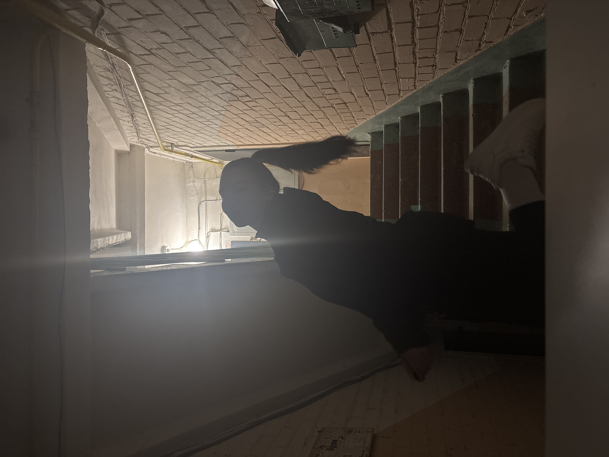 The image depicts a dimly lit interior stairwell within an older multi-story building, characteristic of residential structures in Perm, Russia. A single individual, largely silhouetted, is positioned on the concrete stairs, facing generally upwards and away from the camera. The person, whose gender is not definitively discernible, has long, dark hair styled in a ponytail that hangs down their back, visible against the illuminated background. They appear to be wearing dark clothing. Their right foot, clad in a white sneaker, rests on one of the lower stair treads. The left side of the scene is dominated by a shadowed wall, possibly plastered, with a partially visible white document or sign attached. The right side features an exposed brick wall, painted light brown, upon which two dark metal mailboxes with Cyrillic script are mounted. A yellow gas pipe runs horizontally along this brick wall, alongside exposed electrical conduits and wiring. A small white dome-shaped CCTV camera is visible near the ceiling above the landing, illuminated by an artificial light source, likely an incandescent bulb, which creates a visible lens flare. The concrete stairs feature worn treads and dark-painted risers or stringers, possibly faded green. The overall environment suggests a utilitarian common area, such as a communal hallway or stairwell in an apartment block.