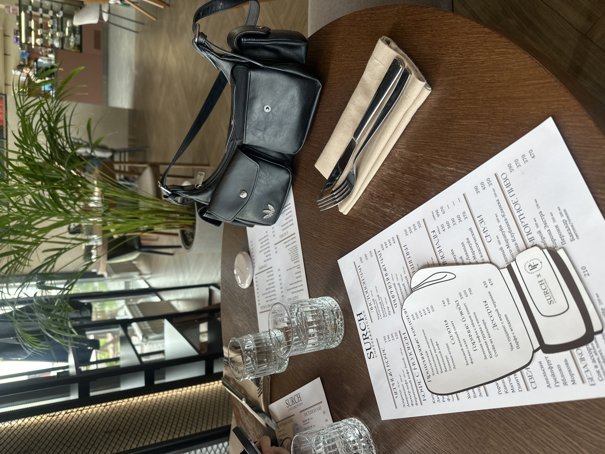 The image depicts a dining table within a cafe or restaurant in Усады, Russia. A round wooden table dominates the foreground, upon which two clear, ridged glasses filled with water are placed. A white menu, printed in Russian with the "SURCH COFFEE & KITCHEN" logo, is spread across the table, partially obscured by a black leather shoulder bag with multiple exterior pockets. One pocket of the bag displays a silver Adidas logo. To the right of the menu and bag, a set of silver cutlery (fork and knife) rests on a folded beige napkin. A small, smooth white object is visible near the top edge of the menu. A hand, likely holding a mobile device, is partially visible on the far left.
The background reveals a brightly lit cafe interior with large windows along the far wall, through which greenery is visible. Various tables and chairs are sparsely arranged, suggesting a spacious layout. A large potted plant with numerous long green fronds stands prominently in the mid-right. In the blurred distance, a counter area with display cases and faint human figures is discernible. On the far left, a black metal shelving unit and a textured screen are visible, with a dark jacket hanging on the shelf. The floor appears to be light-colored concrete or tile.