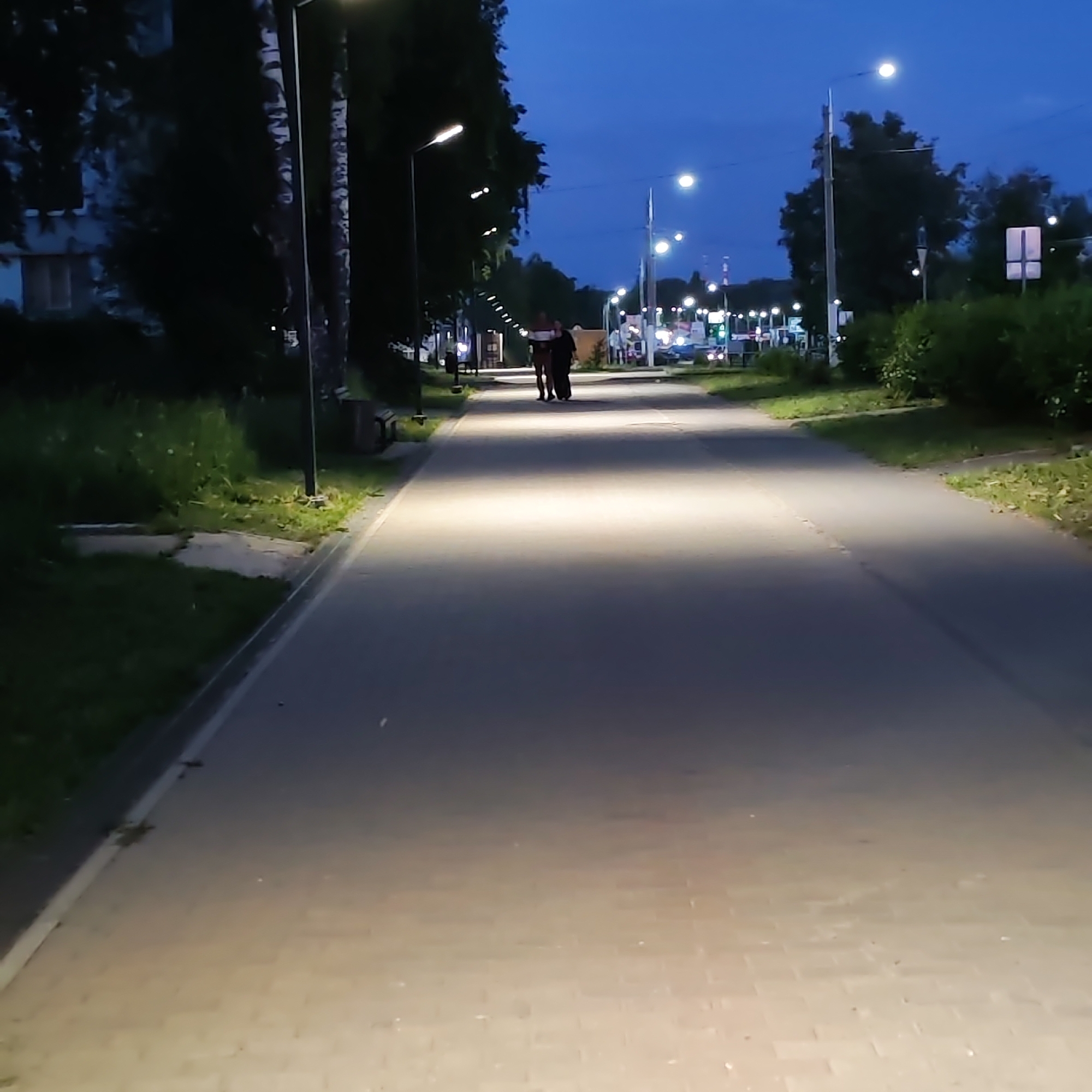 The image captures an outdoor pedestrian scene at dusk or night in Chaikovsky, Russia. A wide, brick-paved pathway extends into the distance, brightly illuminated by a series of modern streetlights. These lights cast long, warm, white beams onto the pathway, creating a well-lit corridor.
In the middle ground, two adult figures are seen walking away from the camera, down the center of the illuminated path. One figure appears to be wearing lighter clothes, while the other is dressed in darker attire. Their silhouettes are discernible against the bright path.
The pathway is flanked by strips of green grass and dark foliage, with tall trees lining both sides. On the left, utility poles with streetlights are visible, and hints of a multi-story building can be seen through the trees. Benches are also present along the left side. The right side features similar vegetation and streetlights, with a more developed urban area, including distant road lights and potential commercial establishments, visible further along the path.
The sky is a clear, deep blue, indicating either early evening or night. The artificial lighting effectively brightens the path, contrasting with the darker surroundings. No discernible text is visible on any signs or objects within the frame.