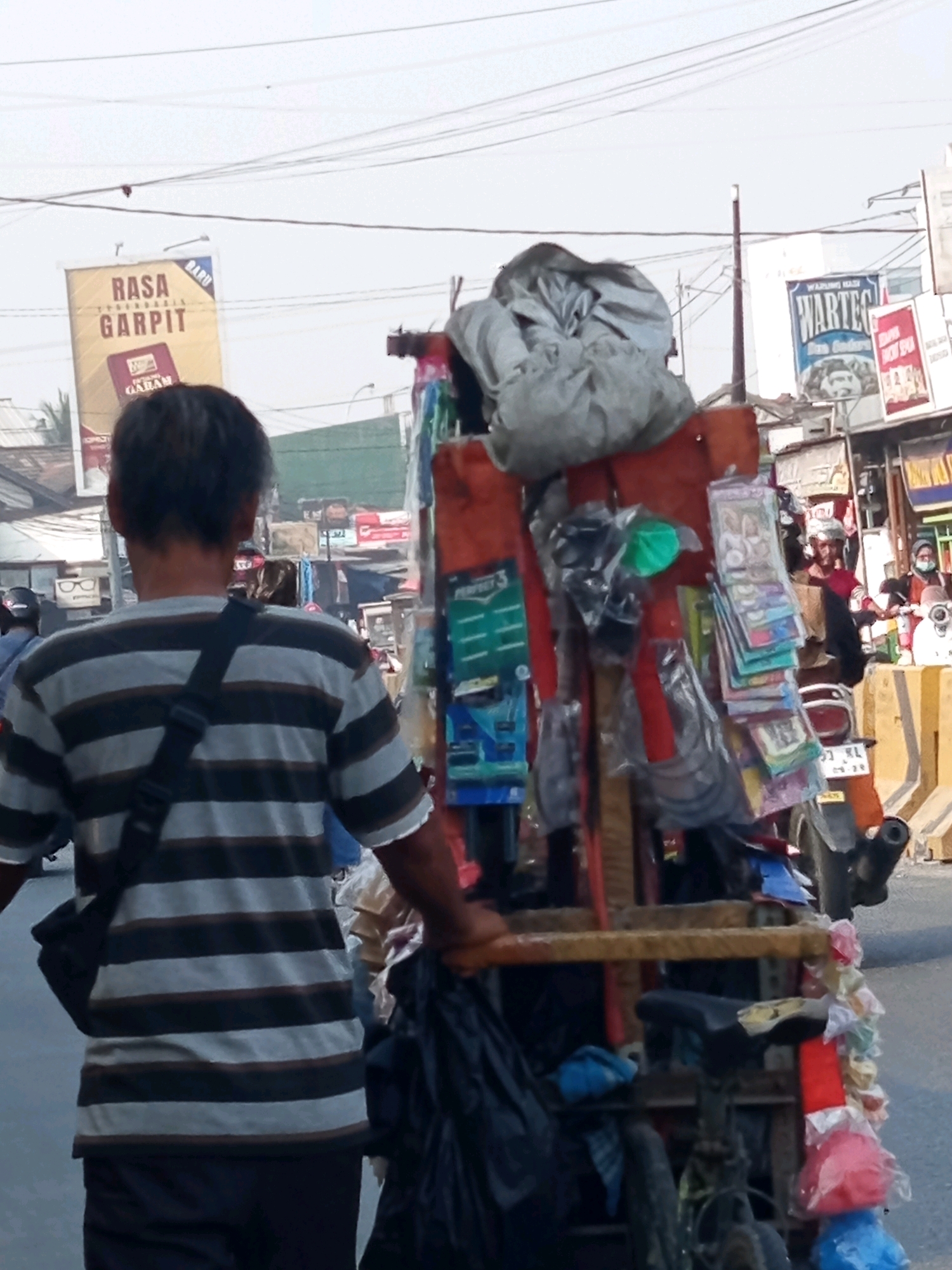 The image shows a street scene in Karawang, Indonesia. In the foreground, a person wearing a striped shirt is pushing a cart filled with various items. The cart is laden with a variety of goods, including what appear to be packaged products, plastic bags, and possibly tools or hardware. In the background, there are buildings with signs, suggesting a commercial area. One sign, visible on the left, reads "RASA GARPIT". Another sign, on the right, reads "WARTEG". Several motorcycles are also present, indicating traffic on the street. The overall environment appears to be an outdoor market or a busy street with shops and vendors. The sky is overcast.