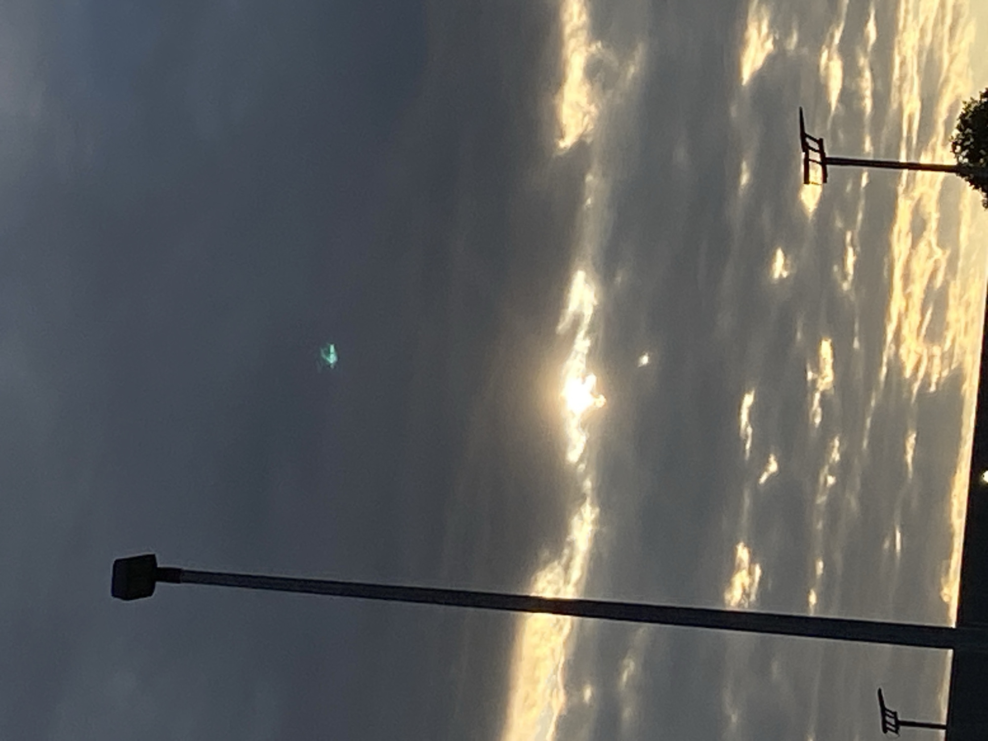 The image shows a cloudy sky with the sun shining through gaps in the clouds, creating a dramatic effect with golden light. A tall, dark lamppost is visible on the left side of the frame, extending from the bottom edge upwards. In the upper right portion of the sky, a small, bright, greenish object is visible. The scene appears to be an outdoor location at dusk or dawn, possibly in the city of Rinteln, Germany, given the context. The overall mood is atmospheric and somewhat mysterious due to the lighting and the presence of the unexplained object.