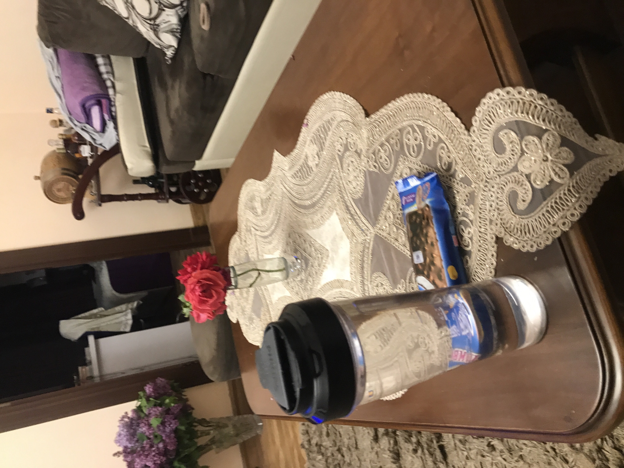 The image depicts a table with a lace runner. On the table, there is a package of Purina Beneful dog treats, a water bottle, and two vases of flowers (lilacs and roses). The table is indoors, possibly in a home. There is a sofa in the background, and a glimpse of an open door. The image does not contain information to identify the location as Yerevan, Armenia.
