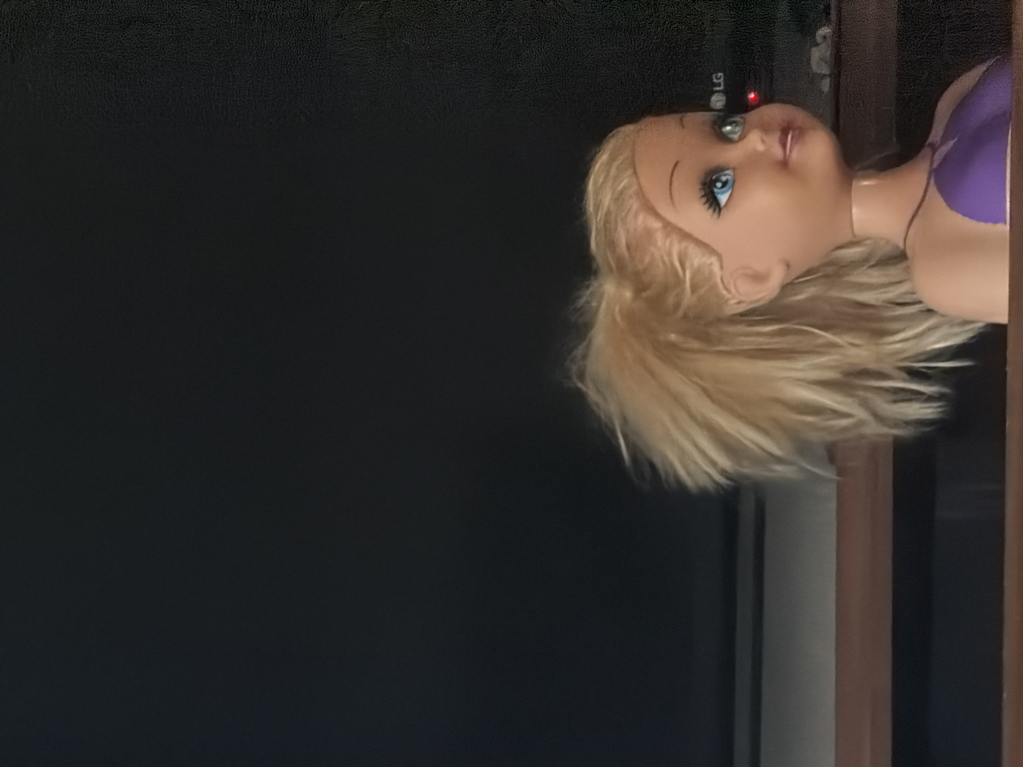 A close-up shot reveals a blonde doll wearing a purple top, positioned on top of a shelf or entertainment center. An LG television is visible, along with other dark-colored furniture. The location of the image is unknown, but is not likely to be in Strasbourg, France.