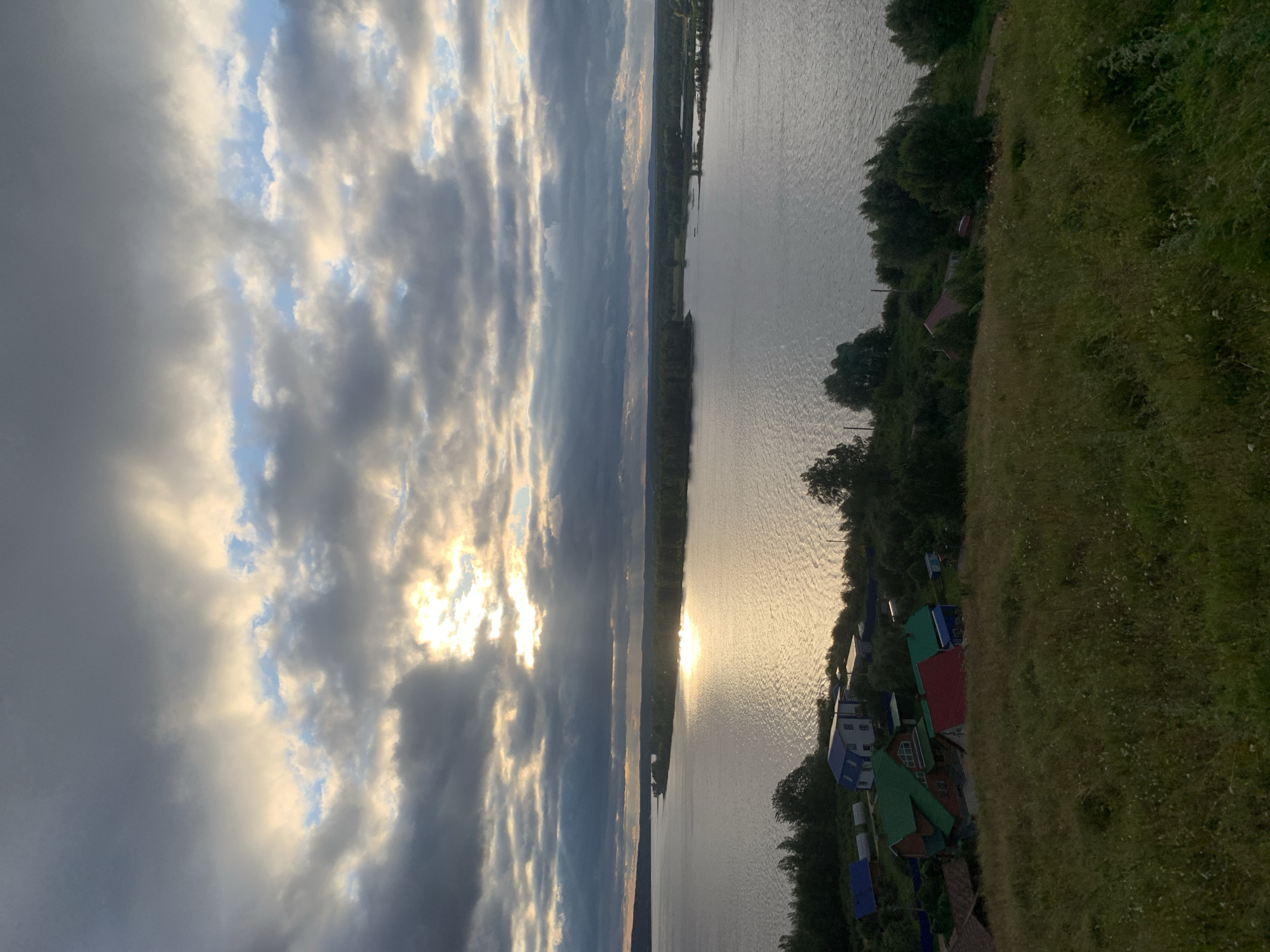 The image captures a serene landscape, likely in the village of Olkhovka, Russia, during what appears to be late afternoon or early evening. A vast body of water, possibly a river or lake, dominates the mid-ground, reflecting the golden hues of the setting sun that is partially obscured by dramatic, heavy clouds. Sunbeams pierce through gaps in the clouds, casting a shimmering light across the water's surface.
The shoreline is fringed with dense green trees and vegetation. On the left bank, a small cluster of houses is visible, characterized by brightly colored roofs, including a prominent red and green-topped structure. These houses suggest a rural or semi-rural settlement. The foreground is an undulating grassy slope, covered in tall, dry grass with scattered small white wildflowers, indicating a natural, uncultivated area. The overall atmosphere is peaceful and somewhat moody, with the interplay of light and shadow creating a visually striking scene. No people or animals are discernible in the image.
