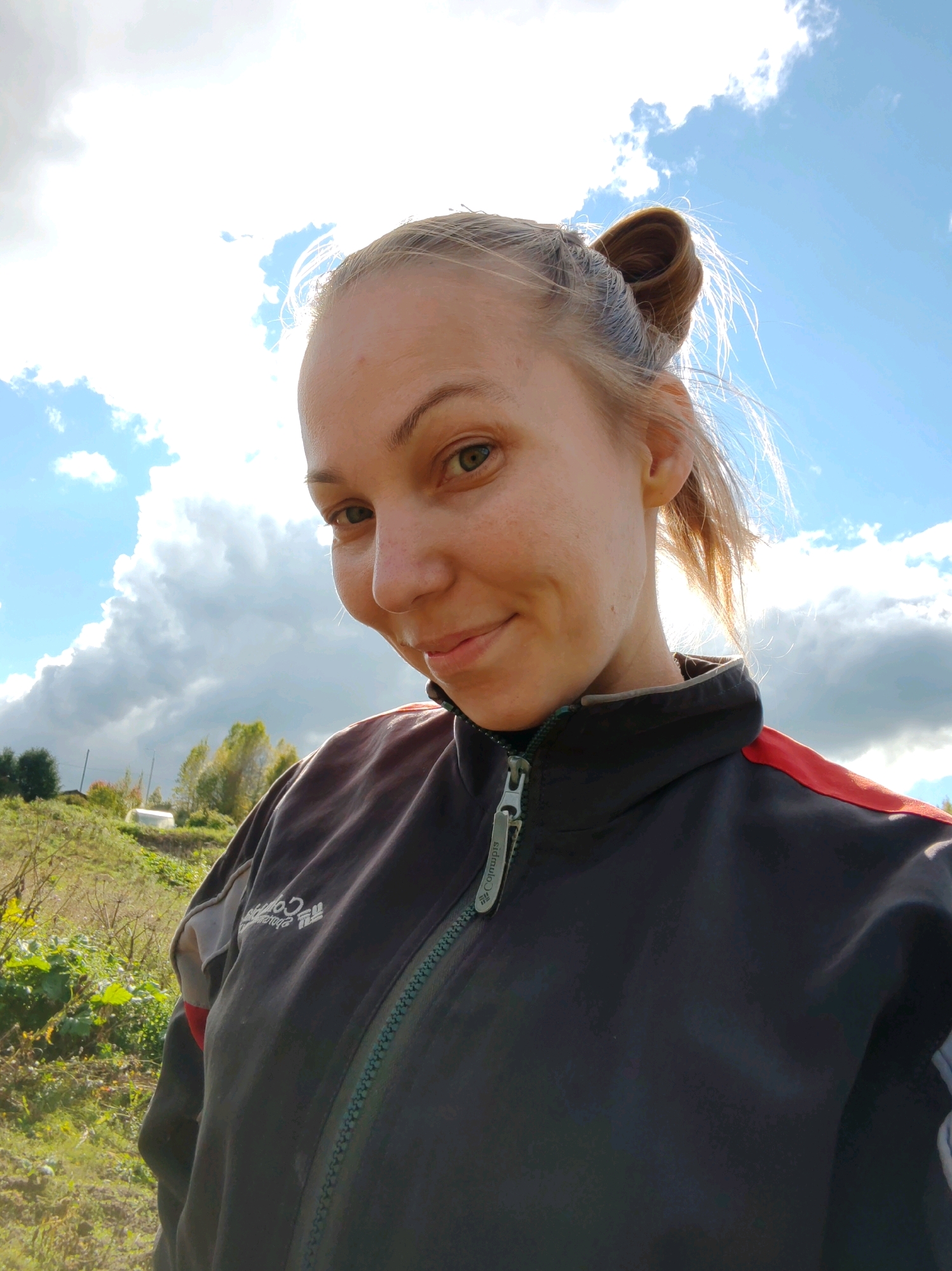A woman with light brown hair pulled into a bun is looking at the camera with a slight smile. She is wearing a dark jacket with red accents on the shoulders and a zipper down the front. The background shows a partly cloudy sky with the sun shining brightly, creating a lens flare. Behind her, there is a grassy hillside with some trees and vegetation. The scene appears to be outdoors, possibly in a rural or natural setting. The location is identified as Chukhlem, Russia.