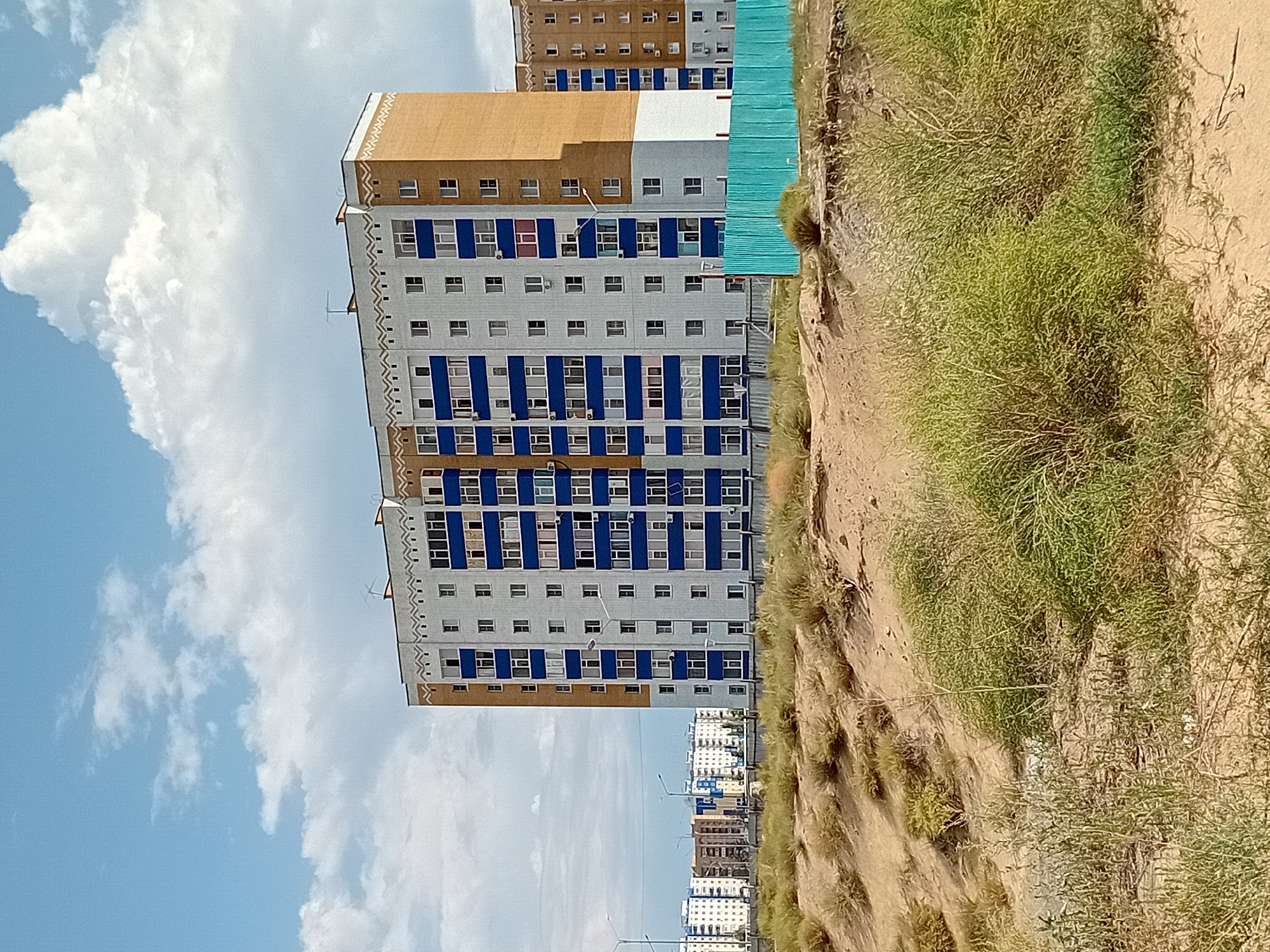 The image shows a multi-story apartment building in the city of Semey, Kazakhstan. The building is light gray with blue accents around the windows and a section of the building is orange. It has multiple windows and balconies. In front of the building is a turquoise fence. The foreground is a sandy area with sparse green vegetation. In the distance, other similar apartment buildings can be seen. The sky is blue with scattered white clouds. The overall scene depicts a residential area.