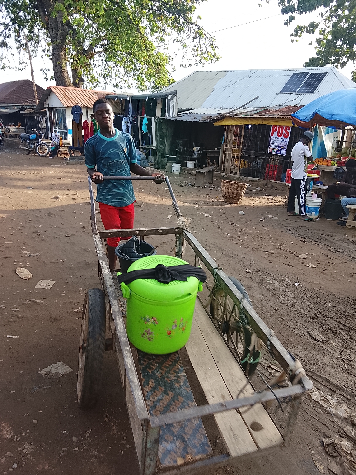 A young man pulls a rickety wooden cart along a dirt road in what appears to be a bustling market area. The cart carries a green bucket with floral designs and a covered black bucket. The man wears a teal and blue patterned shirt and red shorts. He is walking towards the camera.
In the background, there are stalls with merchandise displayed, including clothing. People are milling about, some standing near a fruit stand that features a "POS AVAILABLE HERE" sign. The architecture of the stalls is simple, with corrugated metal roofs. Solar panels are visible on one of the rooftops.
The location is likely in a market area within the city of Abuja, Nigeria, characterized by its busy trade, informal structures, and dirt roads.