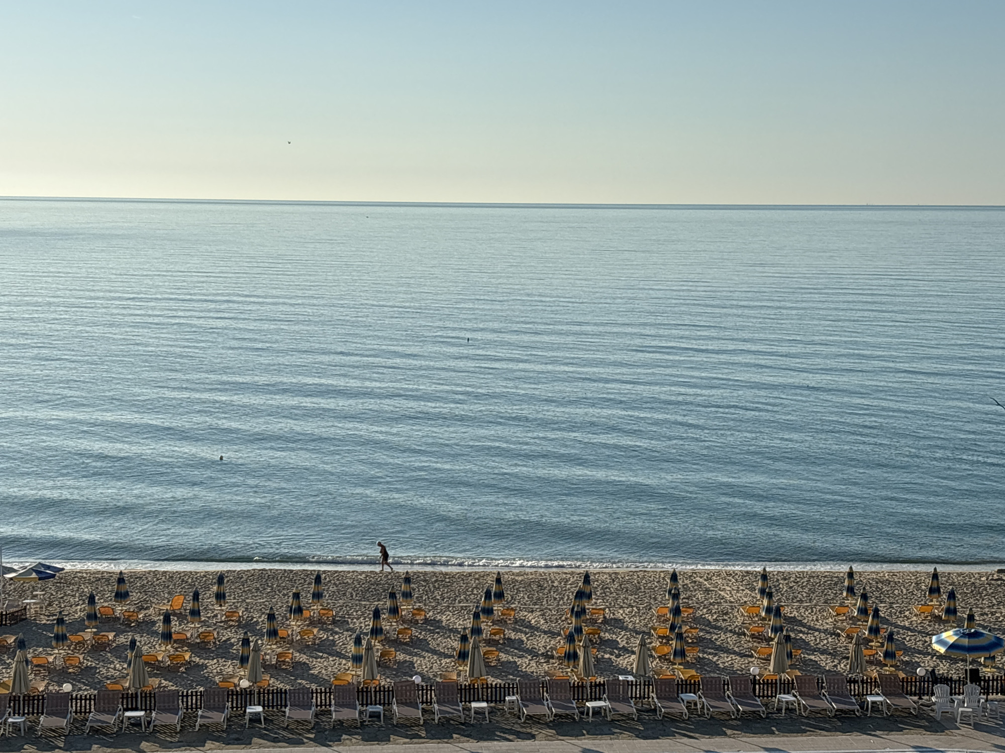 The image depicts a tranquil beach scene with rows of unoccupied lounge chairs and umbrellas set up on the sand. The ocean stretches out to the horizon under a clear, pale blue sky. A lone individual is seen walking along the shoreline where the gentle waves meet the sand. The beach is organized, with a wooden fence separating the lounge area from what appears to be a paved walkway or promenade in the foreground. The scene is bathed in a soft light, suggesting it may be early morning or late afternoon. The location is the beach in Varna, Bulgaria.