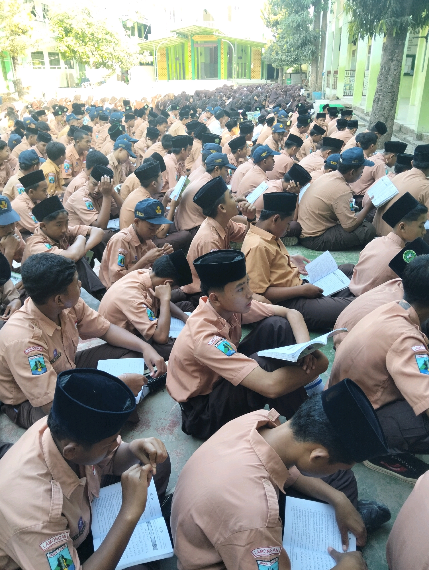 The image depicts a large group of young males, likely students, sitting on the ground outdoors, engaged in reading from open books. They are all wearing similar uniforms consisting of light brown long-sleeved shirts and dark brown pants. Many also wear black peci hats. The students are arranged in rows, filling the foreground and extending into the background. Behind them, a building with green accents and white walls is visible, along with trees and a bright, sunny sky. The ground appears to be a paved or concrete surface. The students' postures suggest a focused and attentive atmosphere, with many looking down at their books. Some students have their hands resting on the books or using them to hold pages open. The overall impression is one of an outdoor educational activity or assembly. The location is identified as Dagan, Indonesia.