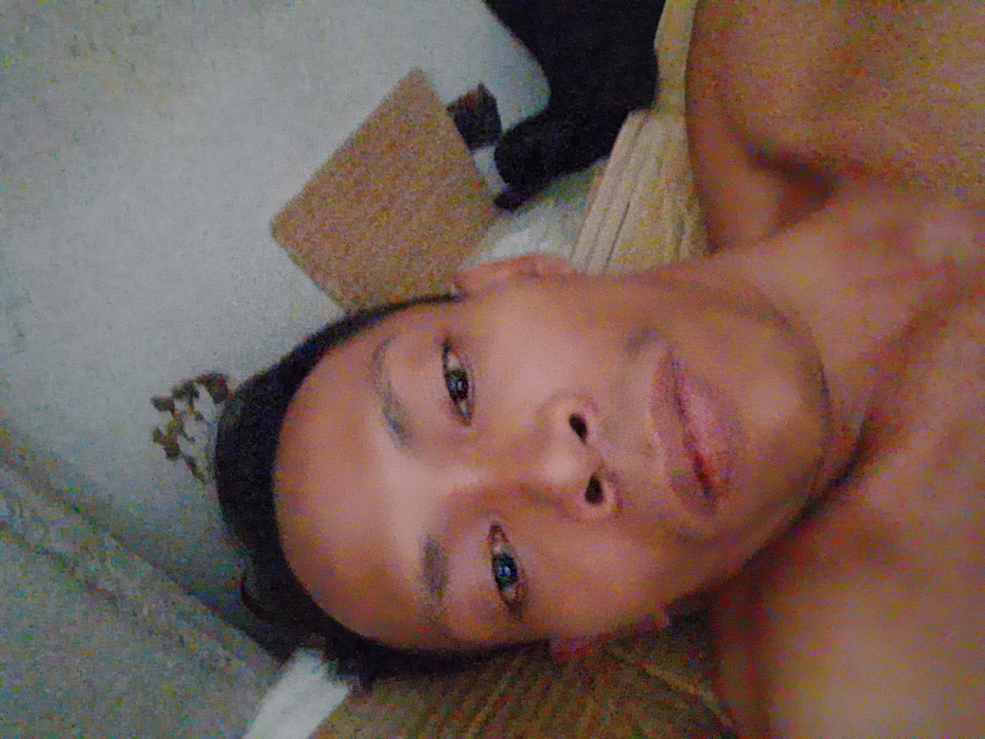 A close-up, slightly angled portrait of a person. The person is facing the camera with their head tilted to the right. The person appears to be lying down on a surface. A light-colored fabric or material is visible underneath the person. The background consists of a wall and a small brown square object. A dark-colored object can also be seen in the background. No specific details are discernible to identify the location as Bukit Ketok, Indonesia.