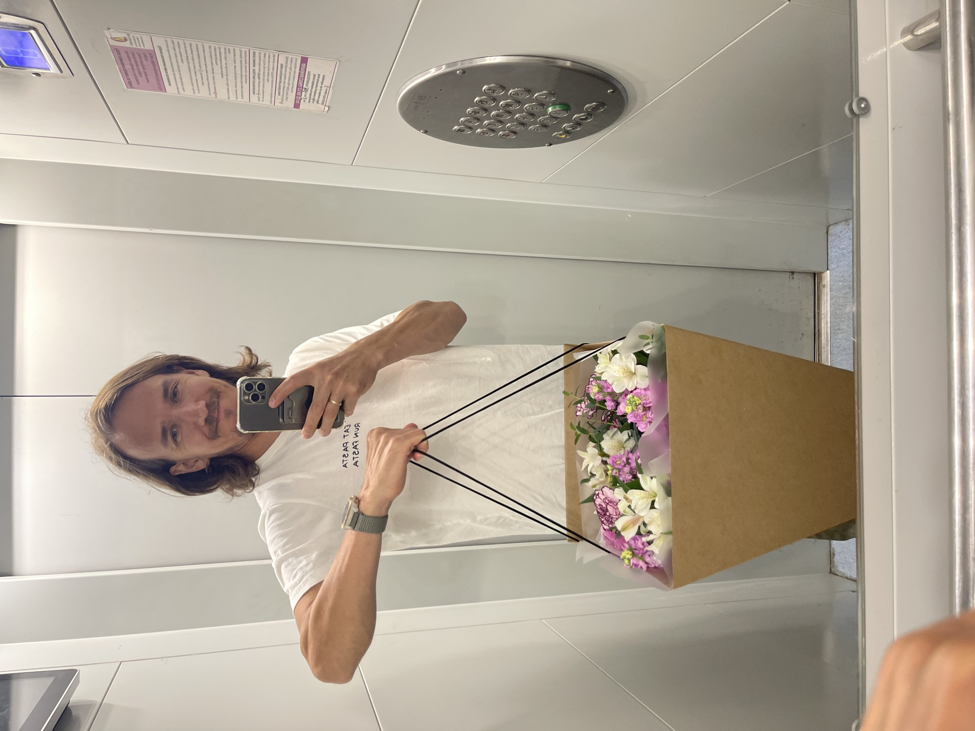 A man with shoulder-length brown hair and a light mustache is taking a selfie in an elevator. He is wearing a white t-shirt with black text and a smartwatch on his left wrist. He is holding a bouquet of pink and white flowers in a brown box with black straps in his right hand. The elevator walls are a light grey color, and there is an elevator control panel on the right side of the image. The floor of the elevator is visible at the bottom of the image.
