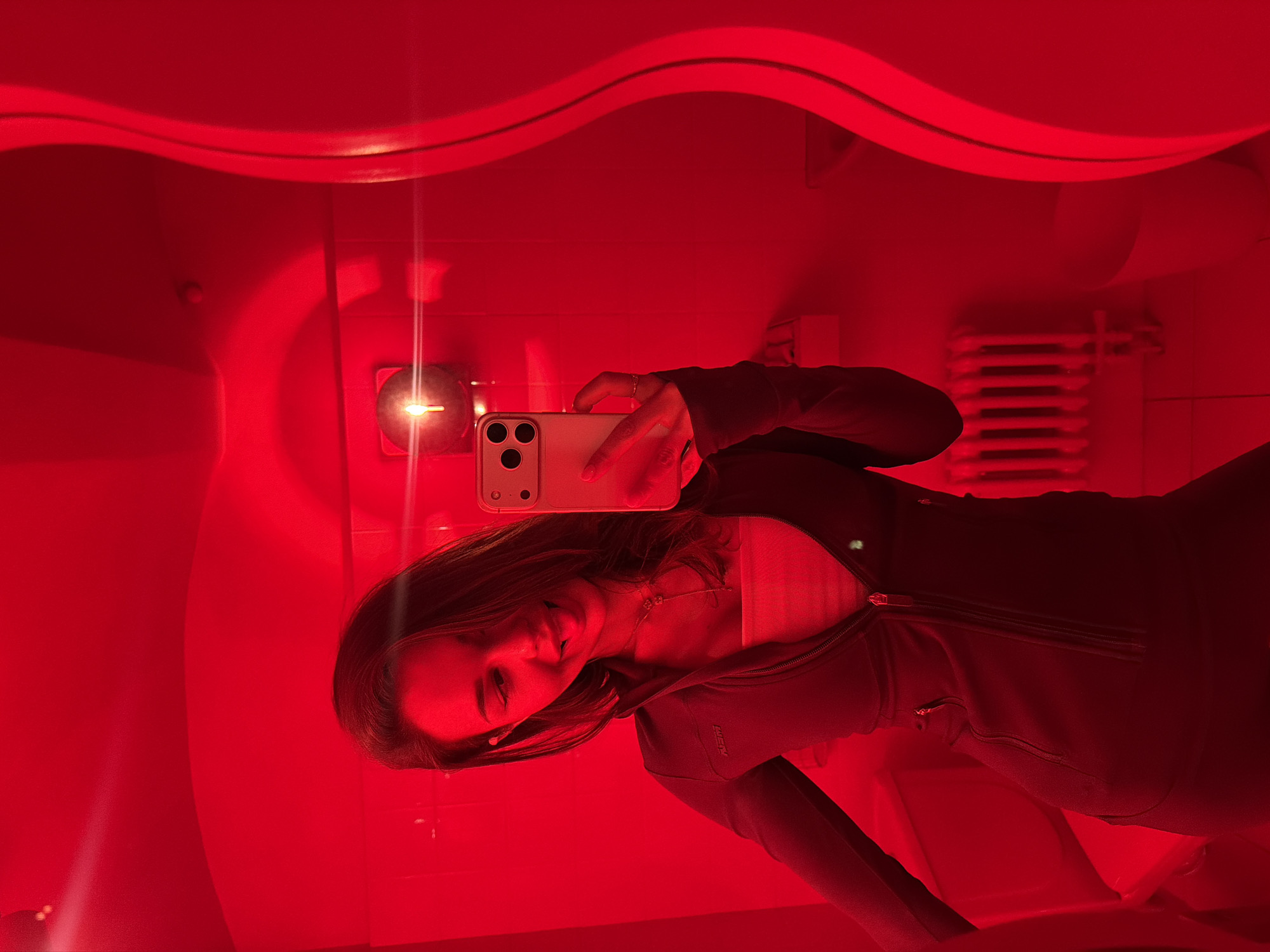 An indoor, mirror selfie features a young woman bathed in intense red light, giving the entire scene a monochromatic crimson hue. The woman, with long dark hair, is seen from the chest up, slightly smiling as she holds a light-colored smartphone (possibly an iPhone) to capture her reflection. She wears a dark, zippered jacket over a lighter top, and a delicate necklace is visible around her neck.
The setting appears to be an enclosed space, potentially a red light therapy room or a uniquely designed interior. Above the woman's head, a distinctive wavy architectural element spans the ceiling. To the viewer's left, a circular light fixture or control panel emits a bright point of light. To the right, behind the woman, a white radiator with horizontal fins is discernible against the red background.
The activity is a self-portrait taken in a mirror. The strong red illumination obscures details of the room but creates a dramatic and intimate atmosphere. No specific time of day is indicated, given the artificial lighting. The woman's expression suggests contentment. There is indistinct text, possibly "NEW" or "NEY", on the left side of her jacket. While the image itself lacks direct visual cues for location, the context provided states Moscow, Russia.