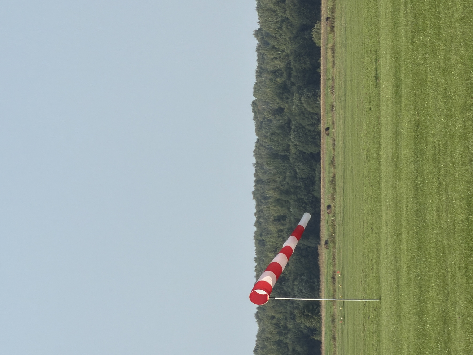 The image depicts an open, grassy field under a clear sky. In the foreground, a windsock, striped red and white, is mounted on a white pole and angled to the left, indicating wind direction. The field is predominantly green, with some patches of dry grass and scattered, dark-colored objects that appear to be hay bales or animal droppings. In the middle ground, a dense line of green trees forms a horizon. The background shows a pale blue, cloudless sky. The scene appears to be in Korobcheevo, Russia, possibly an airfield or a rural open space. There are no people visible in the image.