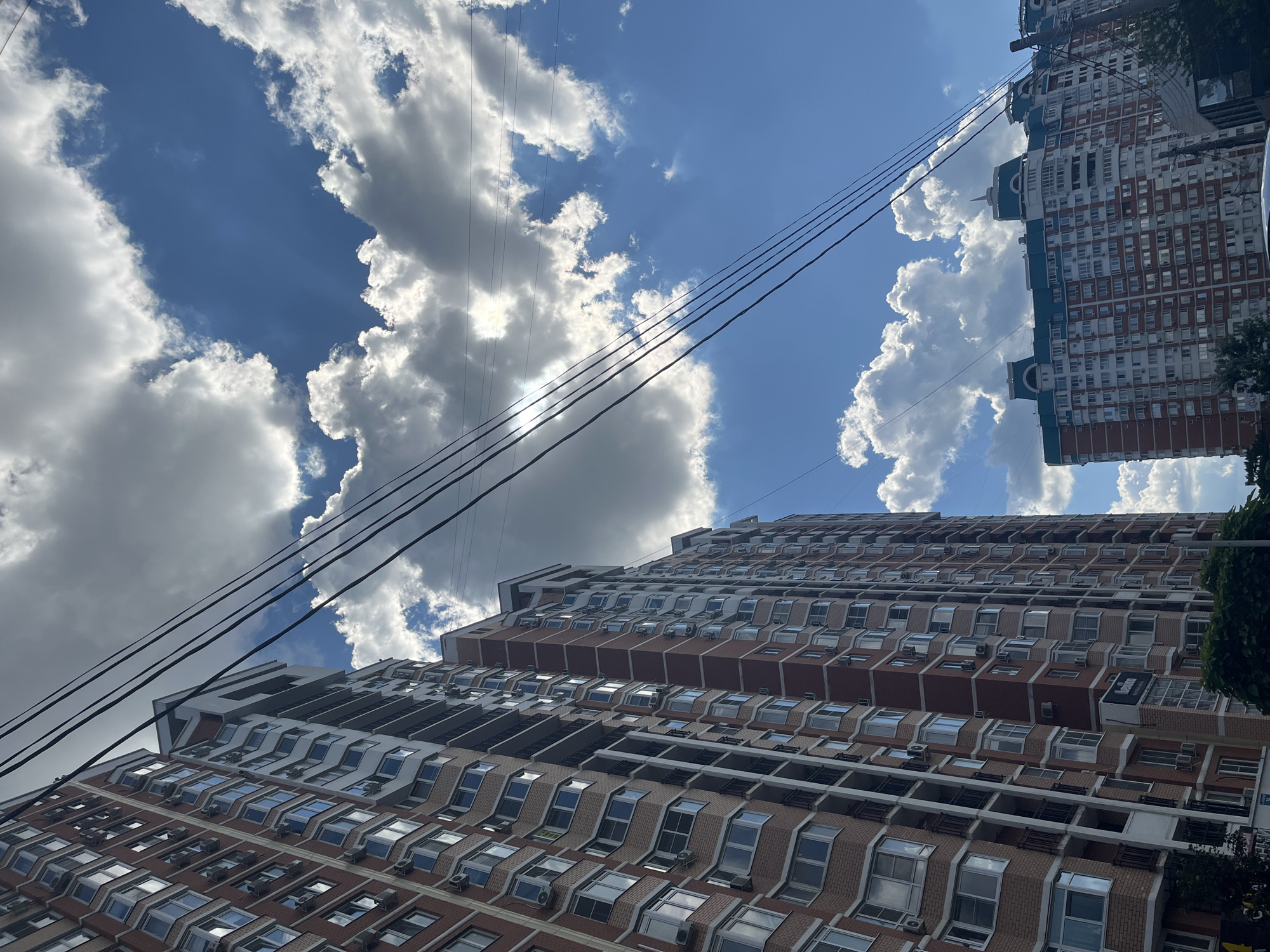 The image depicts an urban scene on a cloudy day, likely in Krasnodar, Russia, based on the architectural style of the buildings and the presence of power lines common in Russian cities. The foreground features a tall, multi-story apartment building with a reddish-brown brick facade and white accents, characterized by numerous windows and balconies. Overhead, thick black power lines stretch across the sky. The sky is a vivid blue, punctuated by large, fluffy white clouds that partially obscure the sun, creating a dappled light effect. In the background, another apartment building, also multi-story, is visible with a distinctive teal-colored roofline. Green trees and foliage are present at the base of the buildings, suggesting a residential area. There are no people visible in the image. The scene conveys a sense of daily urban life, with residential structures dominating the landscape under a dynamic sky.
