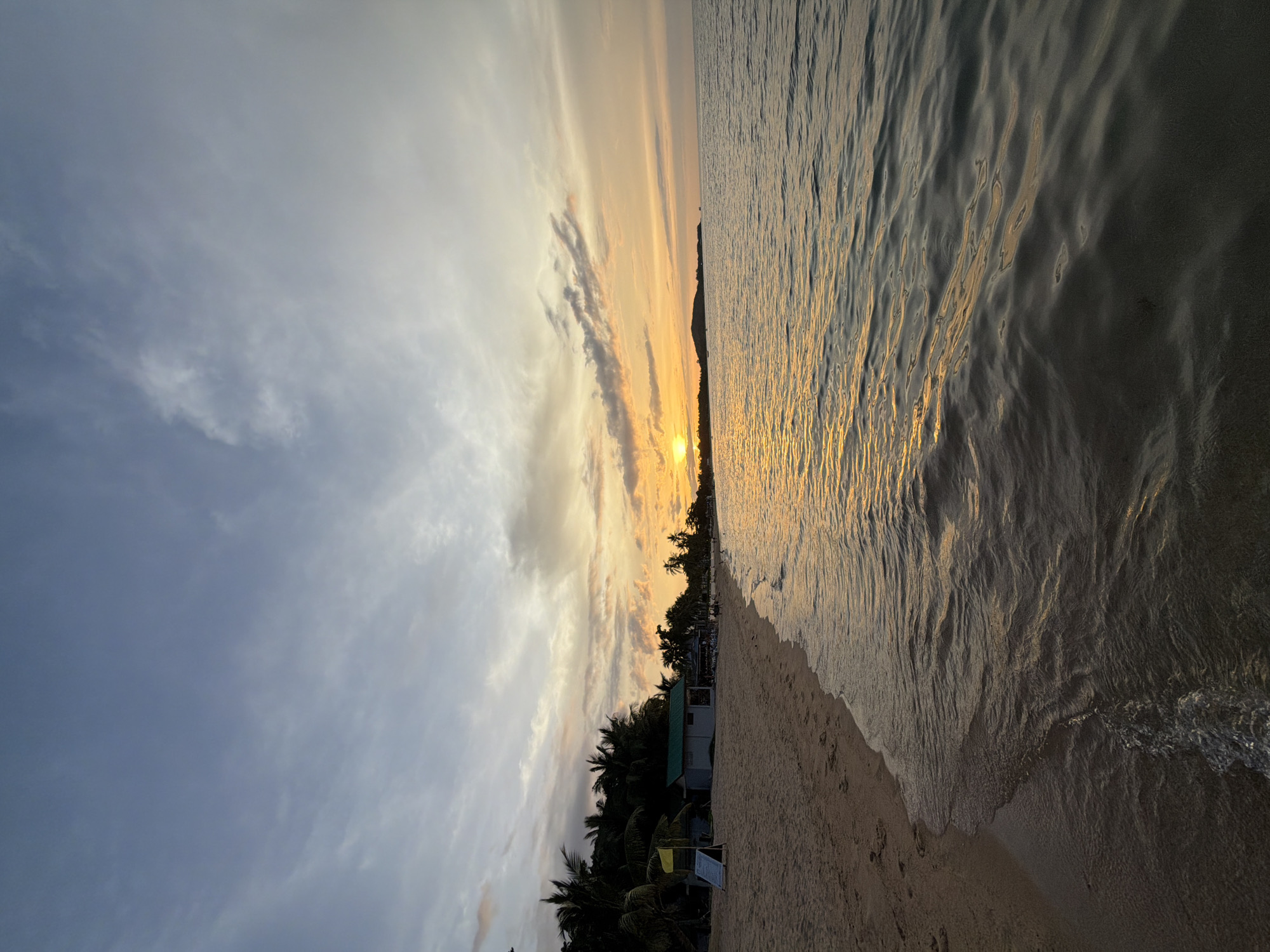 A wide-angle shot captures a beach scene at dusk on Ko Samui District, Thailand. The sun, a bright orange orb, is low on the horizon, casting a warm glow across the sky and reflecting on the calm ocean surface. Wispy clouds streak across the sky, tinged with orange and yellow from the setting sun.
The sandy beach stretches along the left side of the frame, with footprints visible in the sand. A few palm trees line the edge of the beach, their dark silhouettes contrasting with the bright sky. A small building with a green roof and a yellow flag on a stand are also present on the beach. The ocean occupies the right half of the image, with gentle waves lapping at the shore. In the distance, a faint silhouette of a landmass can be seen. No people are clearly visible in the shot, though there may be distant figures on the beach. The overall impression is one of tranquility and natural beauty.