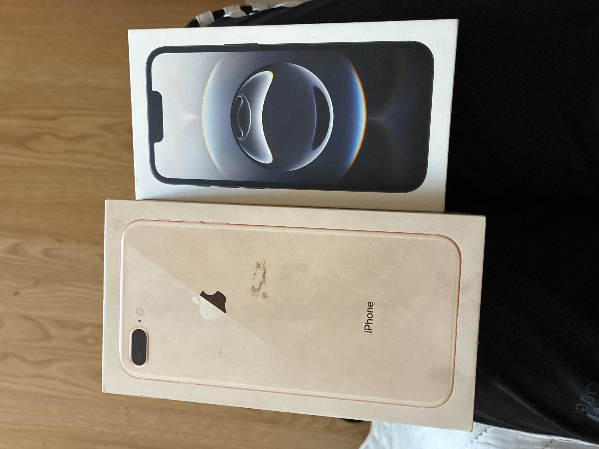 The image displays two iPhone boxes, stacked and slightly offset, resting on a surface that transitions from a light wooden floor or table on the left to a dark, possibly fabric, material on the right.
The top box is white and features a prominent, full-screen depiction of a modern iPhone, likely a Pro model, with its screen displaying an abstract, dark wallpaper with luminous circular elements and colorful, iridescent reflections. The distinctive notch of the iPhone is visible at the top of the depicted screen.
Beneath it, the bottom box is a light gold or rose gold color. This box showcases the back of an older iPhone model, identifiable as an iPhone 8 Plus due to its horizontal dual camera array and the classic Apple logo positioned centrally. The surface of this box appears somewhat worn, with noticeable smudges and dirt marks, particularly above and to the right of the Apple logo. The word "iPhone" is printed vertically on the right side of this box.
The setting appears to be indoors, with the lighting suggesting an ambient, perhaps daylight, environment, though no specific time of day can be determined. The image focuses solely on these objects, with no people or discernible activity taking place.