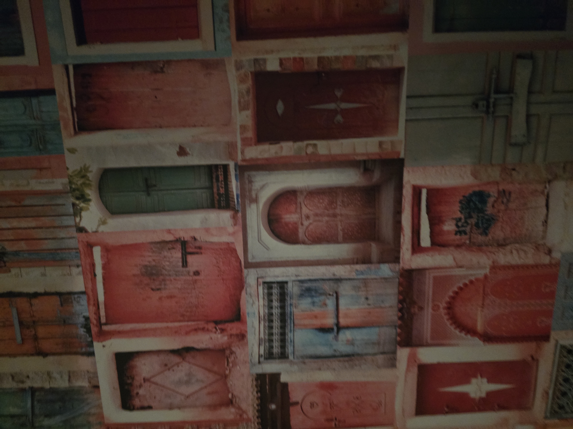 This media file presents a collage or pattern composed of numerous stylized doors, arranged in a grid-like, vertical composition. The overall impression is that of a decorative surface, possibly wallpaper or a mural, rather than an actual physical scene.
The subjects are exclusively doors of various shapes, sizes, and colors, often appearing aged, rustic, and weathered. Key details include:
- **Colors:** A dominant palette of reds, oranges, and browns, interspersed with muted greens and blues. These colors often appear faded, distressed, or with peeling paint effects.
- **Designs:** The doors showcase diverse architectural and decorative styles. Some are simple paneled doors, while others feature intricate carvings or metalwork. Notable designs include:
- An arched door in the central area with elaborate relief patterns.
- A reddish-brown door (top center) with a white diamond shape and a star-like motif.
- Another red door (bottom right) with a prominent white starburst or cross design.
- A red door (mid-right) adorned with a stylized blue motif resembling a tree or floral pattern.
- Visible hardware such as handles, hinges, and latches adds to the antique aesthetic.
- **Surroundings:** The areas between and around the doors are depicted as distressed wall sections, showing textures of brick, plaster, or peeling paint, reinforcing the aged look. A small amount of green foliage is visible near one green door.
There is no activity or event taking place; the image is a static representation of architectural elements. The aesthetic evokes a sense of history, charm, and cultural heritage, aligning with the architectural character often found in historical cities such as Plovdiv, Bulgaria. No discernible text is visible anywhere in the image.