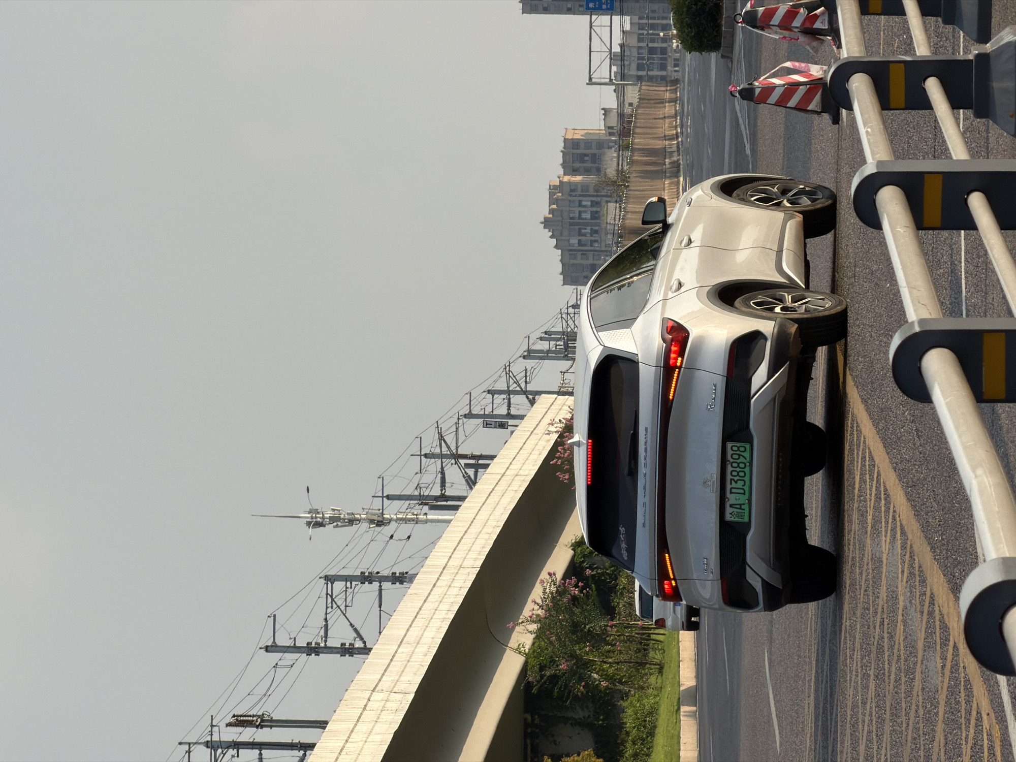 In the city of Jiangjin, China, a white BYD Tang DM-i SUV is captured from behind, driving on a multi-lane road. The vehicle is in the right lane, with its brake lights illuminated. The license plate is clearly visible as "A D38898". To the left of the SUV, a white Volkswagen Polo is visible in the adjacent lane. Overhead, a concrete overpass, a common feature in Chinese cities, spans across the road, supported by pylons. Complex overhead electrical wires, typical of a railway or power grid infrastructure, are strung across the sky. The environment appears urban, with buildings visible in the background. Construction barriers with red and white stripes are placed on the roadside, indicating ongoing work or lane closures. The sun is low in the sky, casting a warm light on the scene.
