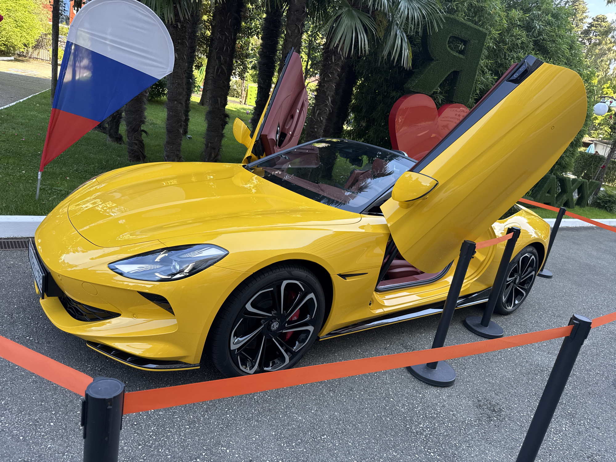 An eye-level, medium shot captures a bright yellow sports car with its doors open, parked on an asphalt surface outdoors. The car's interior appears to be a dark red. A Russian flag is visible in the background to the left, and tall palm trees line the right side of the frame. In the distance, large, green, hedge-like letters spelling out "Я" (Ya, meaning "I" in Russian) and "AXI" are prominent. Red heart decorations are also visible behind the car. An orange safety barrier cordons off the area in front of the car. The scene is likely set in Sochi, Russia, given the presence of the Russian flag and the characteristic vegetation.