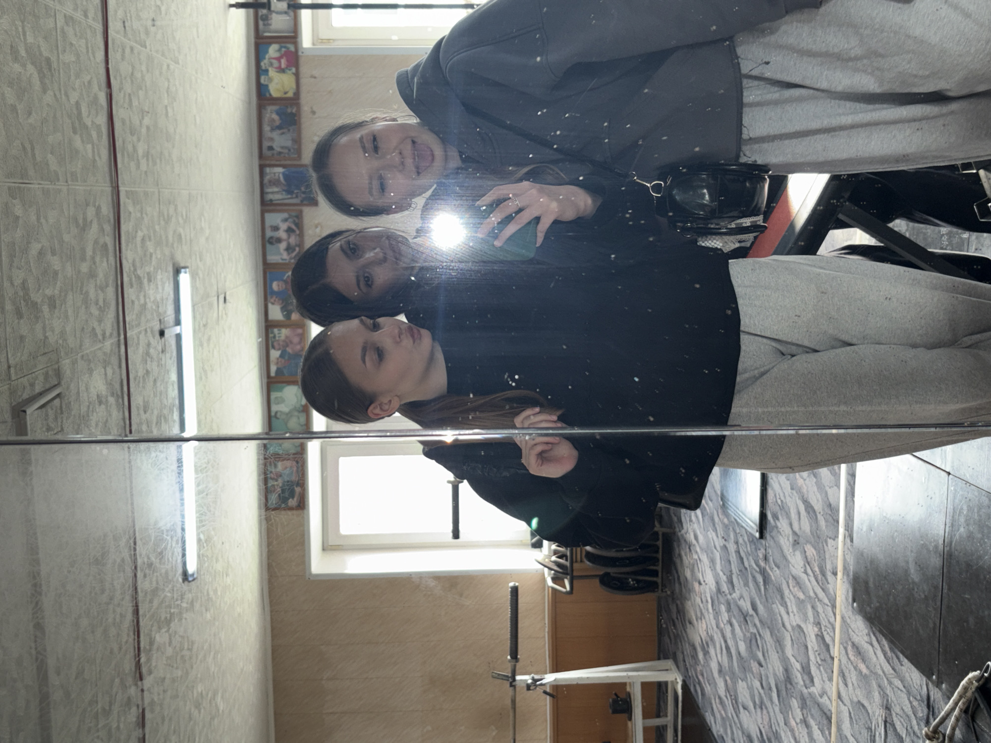 Three young women are taking a mirror selfie in a gym. The woman on the left is pouting, and the woman on the right is sticking her tongue out. They are both wearing grey sweatpants and black hoodies. The woman in the middle is looking at the camera. The gym appears to be older, with wood-paneled walls and fluorescent lighting. There are some framed pictures of men on the wall, possibly bodybuilders. Weights and gym equipment are visible in the background. The floor is carpeted with a grey, abstract pattern. The reflection in the mirror shows the gym in more detail, including a window, a barbell, and a rack of weights. The image is lit by the flash of the phone, creating a bright spot in the center of the frame.