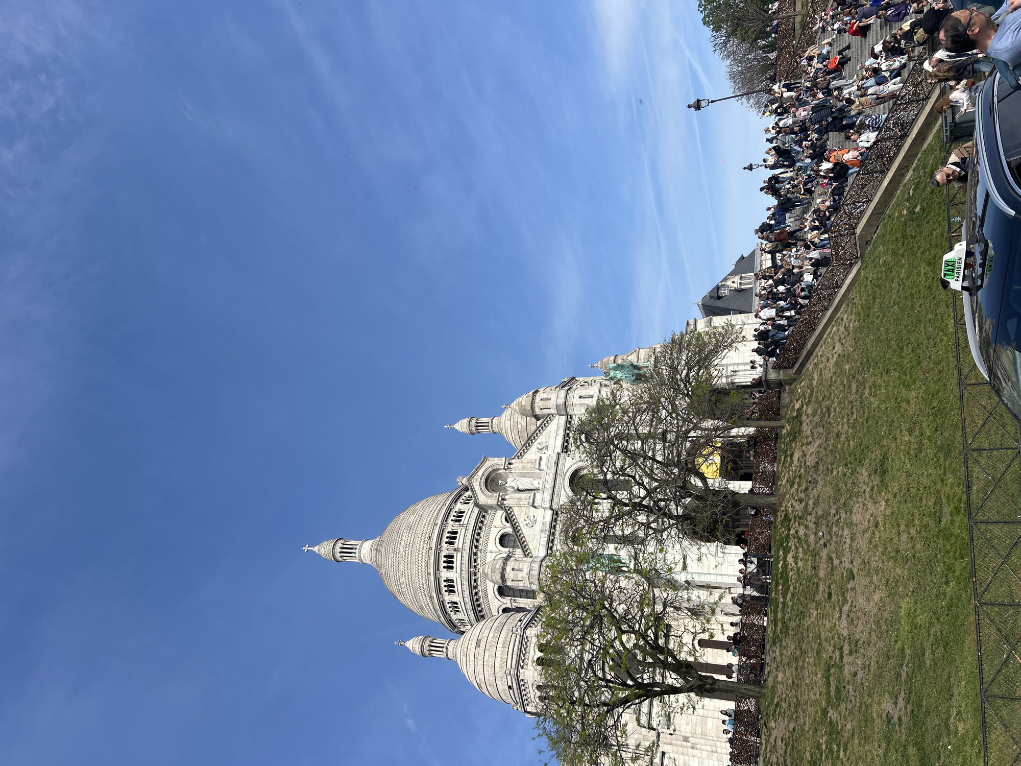 The image is of the Sacré-Cœur Basilica in Paris, France. The basilica, a large white stone building with multiple domes, is the primary subject. Trees with few leaves are in the foreground and partially obscure the lower sections of the building. In the background and to the right, people sit on steps. A dark-colored car, identified as a taxi by a sign on its roof, is partially visible on the right edge of the image. There is a clear blue sky.