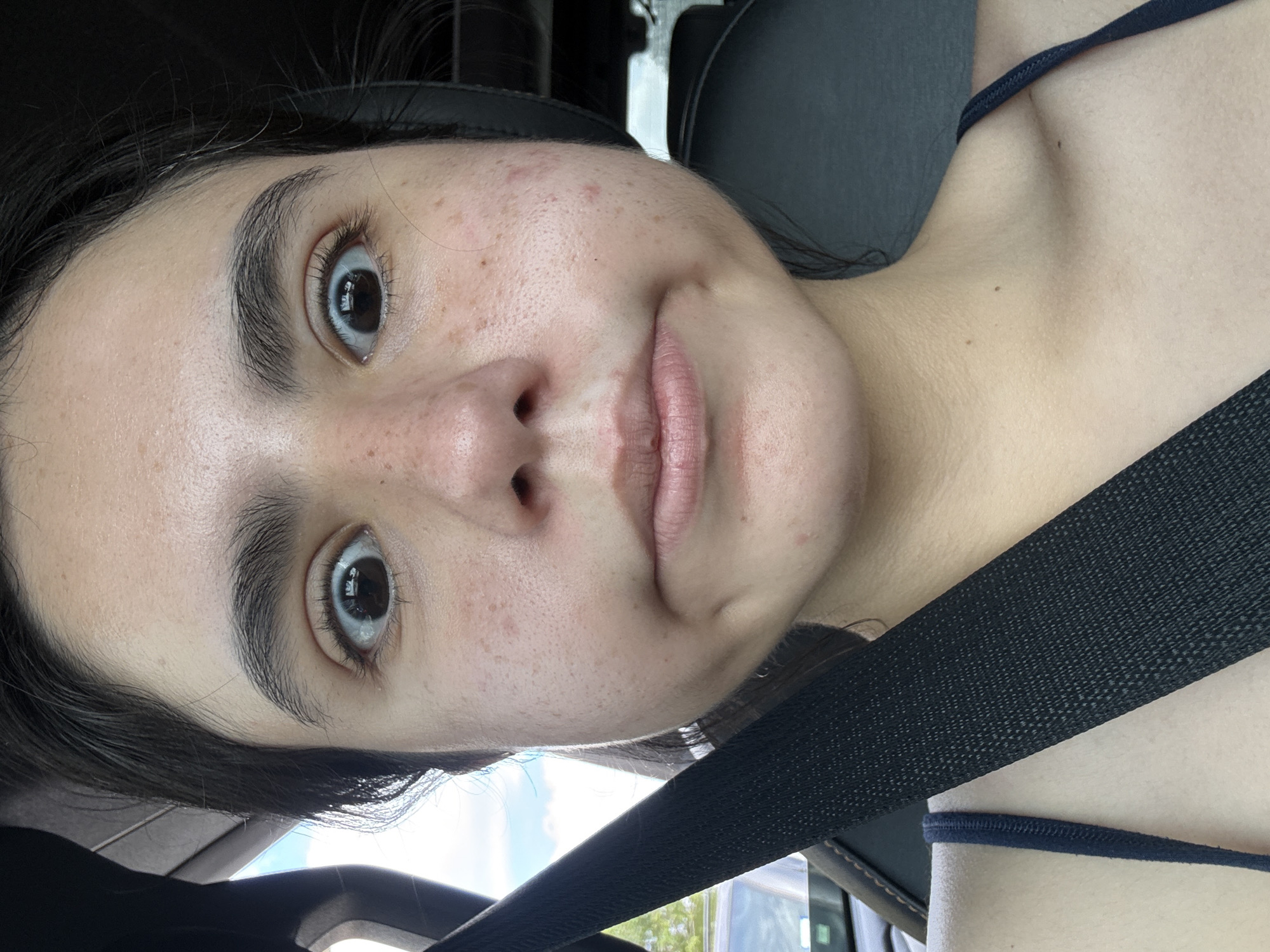 A close-up selfie of a young woman, presumably in her late teens or early twenties, is the primary subject of the image. She is positioned in what appears to be the driver's seat of a car, as evidenced by the black seatbelt diagonally across her body and the car interior visible in the background. The camera angle is slightly below her, tilting upwards towards her face. Her gaze is directed towards the camera with a slightly surprised expression, as evidenced by her wide eyes. She has fair skin, dark hair, and noticeable freckles and some acne on her face. The lighting suggests an indoor environment with natural light filtering through the car windows. The specific location within the city of Austin, United States of America, cannot be determined from this image alone, as there are no identifiable landmarks or street signs visible.