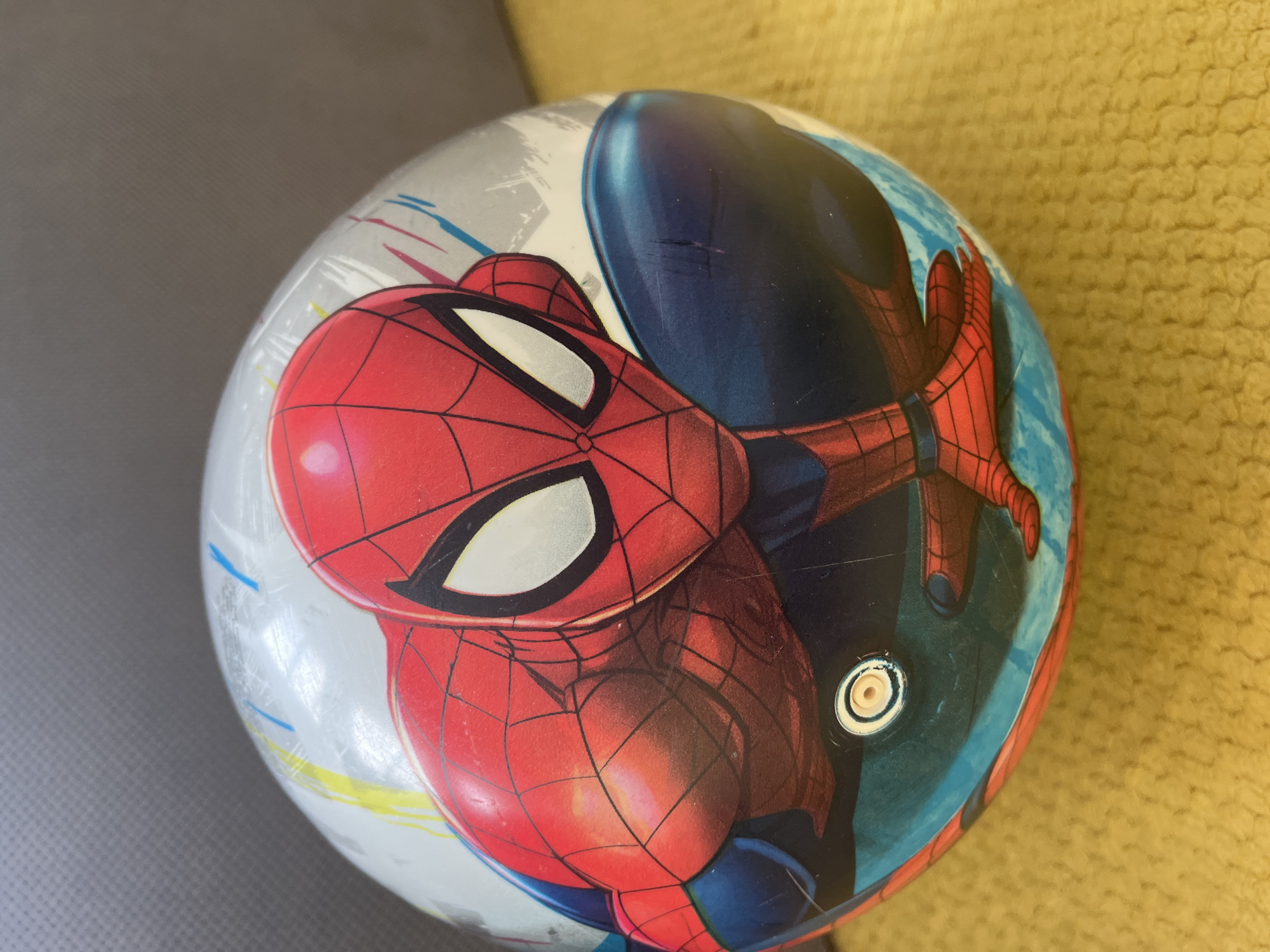 A Spiderman ball sits on a surface with a gray and yellow portion. The ball features the Spiderman character in his signature red and blue suit. The location is unknown.