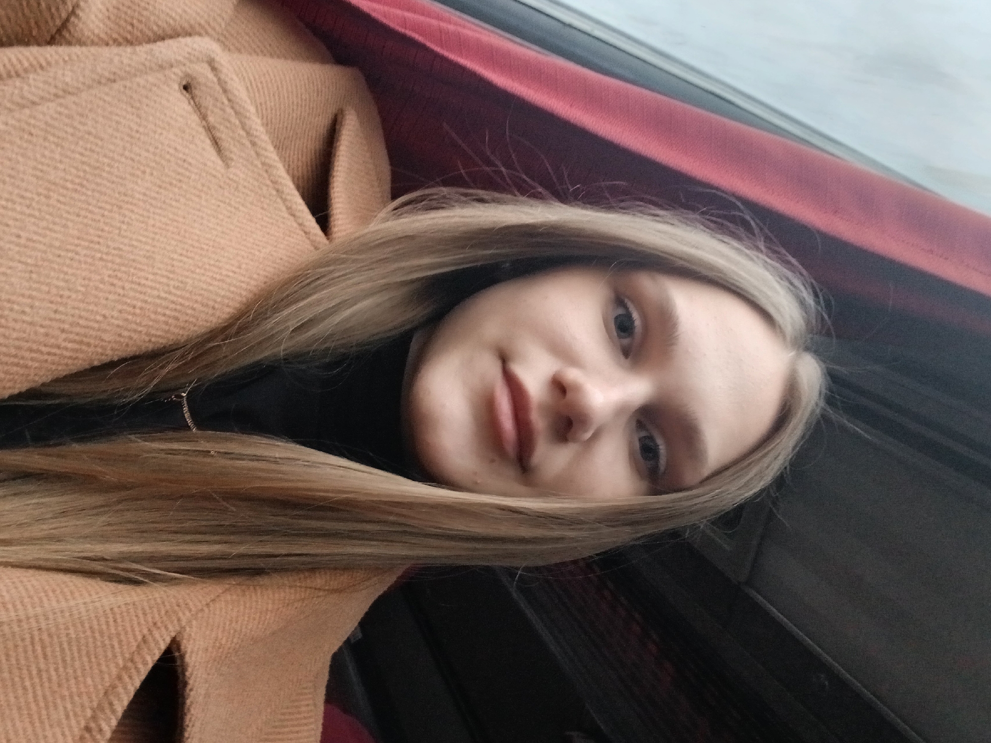 A young woman with long blonde hair and light skin is shown in a selfie-style photograph. She wears a black turtleneck sweater and a tan overcoat. The background suggests she is inside a vehicle; a maroon surface is visible behind her head, and a window is to her right. The photograph is oriented at a slight angle. No specific details are discernible to confirm the location as Chernovskoe, Russia. The photo solely shows a young woman inside a vehicle.