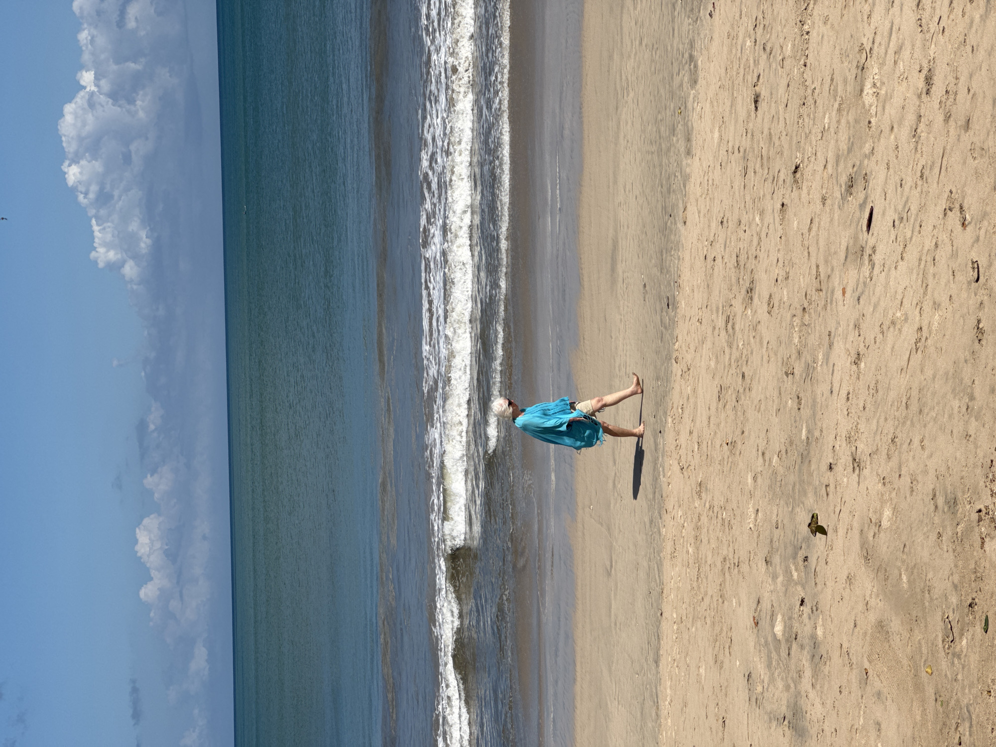 A middle-aged person is walking barefoot on a sandy beach toward the right. The person is wearing a turquoise or blue coverup and tan shorts. The person has gray hair. To the left of the person are ocean waves and a blue sky with puffy clouds. The beach sand is light tan in color. A dark shadow is cast to the right of the person. A leaf is visible on the right. The location may be Kuta Selatan, Indonesia, but there is no verifiable evidence in the image.