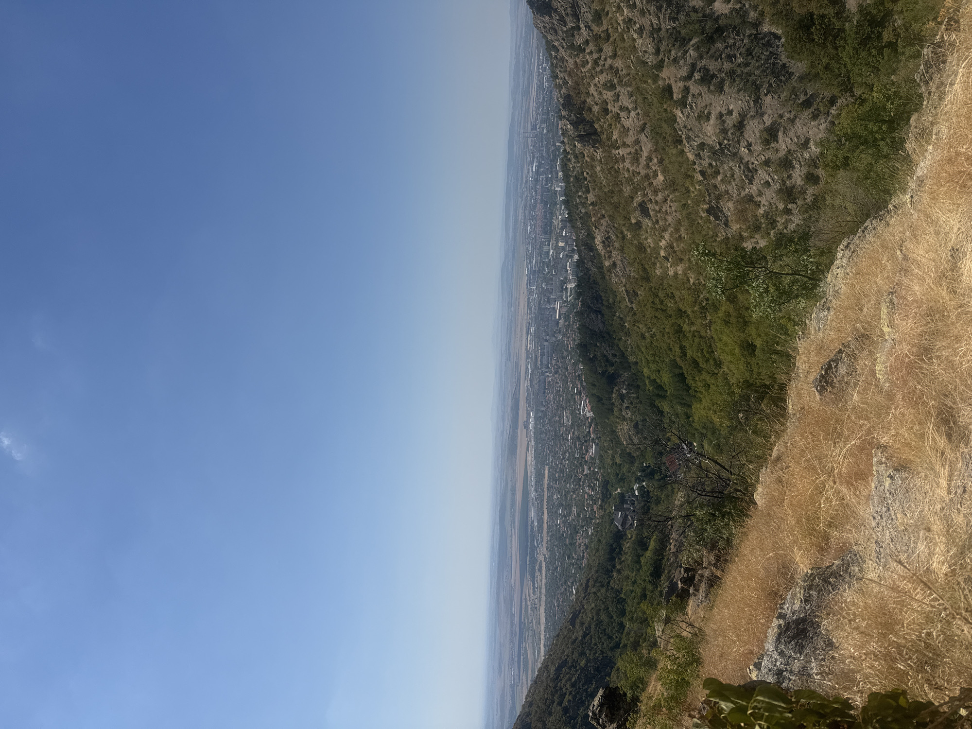 The image is a wide-angle, high-angle shot overlooking the city of Sliven, Bulgaria, from a rocky, elevated vantage point. The foreground features dry, golden grass and rocks, typical of a hillside in warm weather. A sparse green plant is visible on the left edge. Descending into the middle ground, a valley is filled with dense green trees, with a few buildings nestled within. The middle distance shows a sprawling urban area with numerous buildings, including some taller structures and what appear to be industrial facilities or power plants with smokestacks in the far distance. The background consists of a hazy, flat plain that extends to the horizon under a clear, pale blue sky. The overall impression is of a city nestled within a valley, viewed from a natural overlook on a sunny day. There are no people or animals visible in the image.