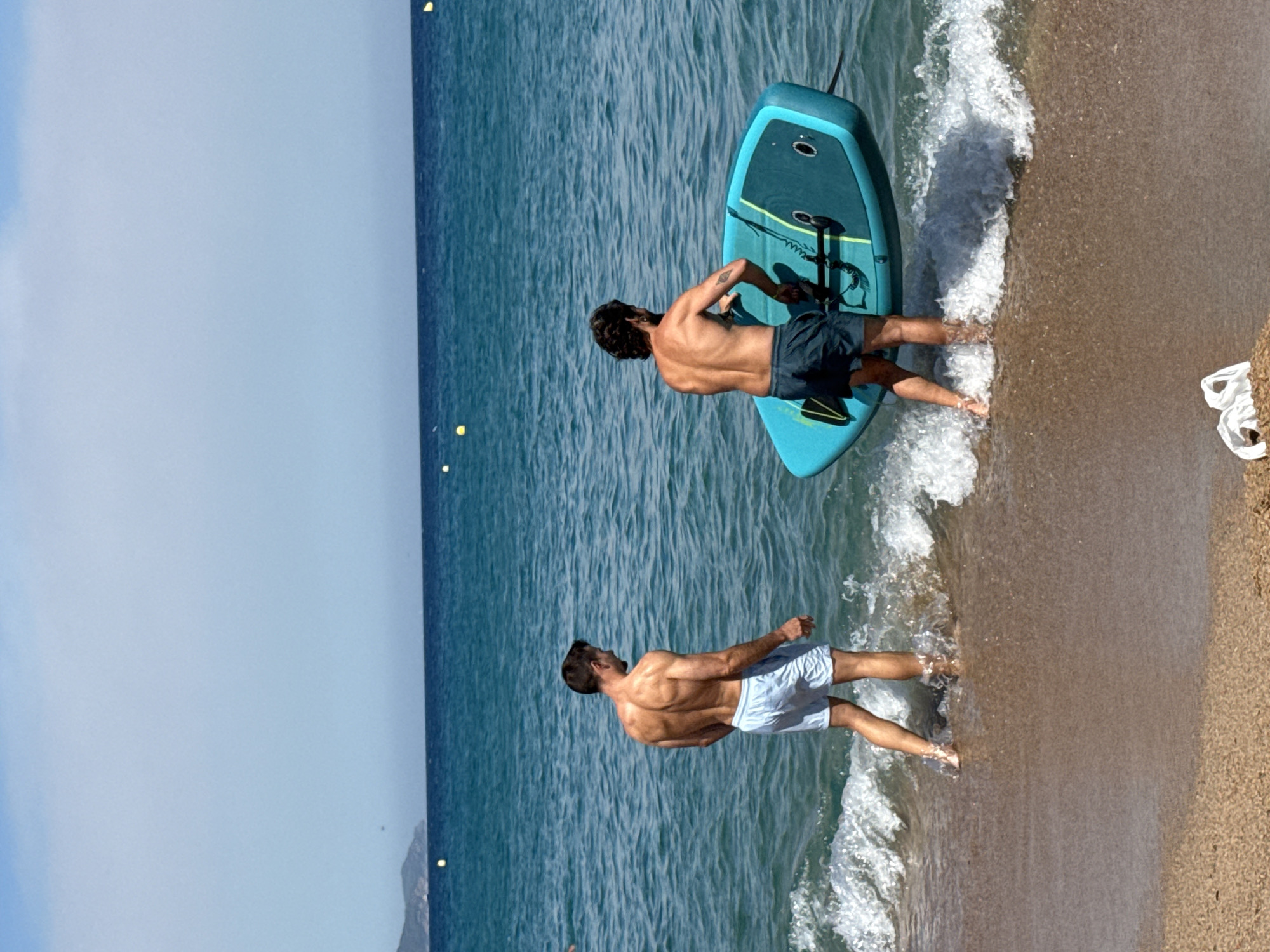 Two men stand in the shallow water of a beach in Blanes, Spain. The man on the left, facing away from the camera, wears light blue swim shorts and has a muscular build. The man on the right, also facing away, wears dark swim shorts and carries a teal paddleboard with yellow accents. The paddleboard is partially submerged in the turquoise water, and a black leash is attached to it. Gentle waves are breaking on the sandy shore. In the background, the Mediterranean Sea stretches to the horizon, with a hazy coastline visible in the distance, including a hill with some greenery. The sky is a clear, pale blue with some wispy clouds. The lighting suggests it is daytime, likely during the summer months. A small white object, possibly trash, lies on the sand in the foreground.